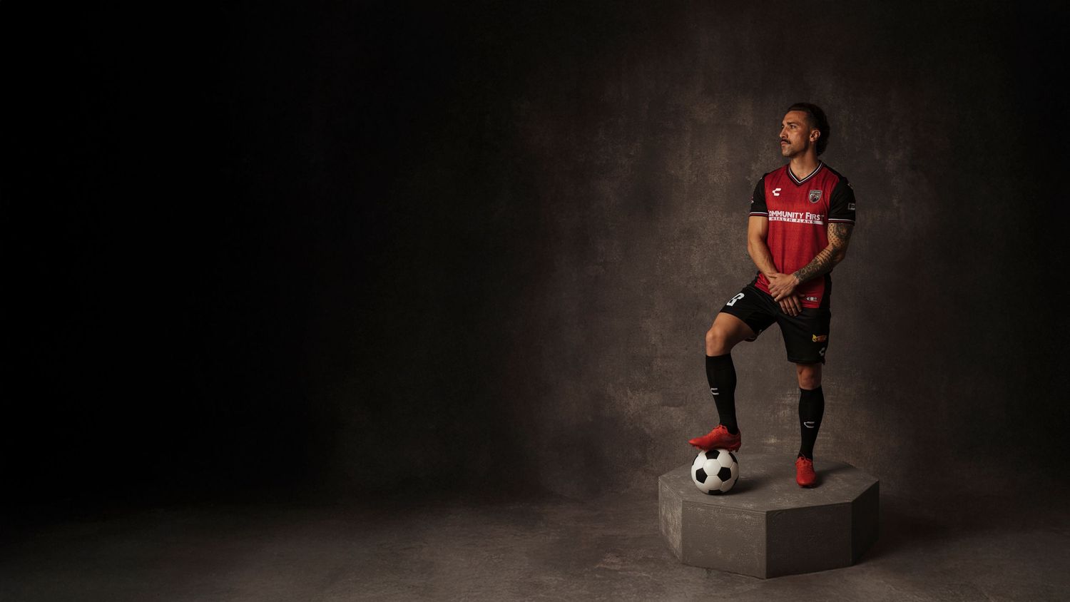 Soccer player in red and black uniform stands on a platform with a soccer ball in a dramatic studio setting.