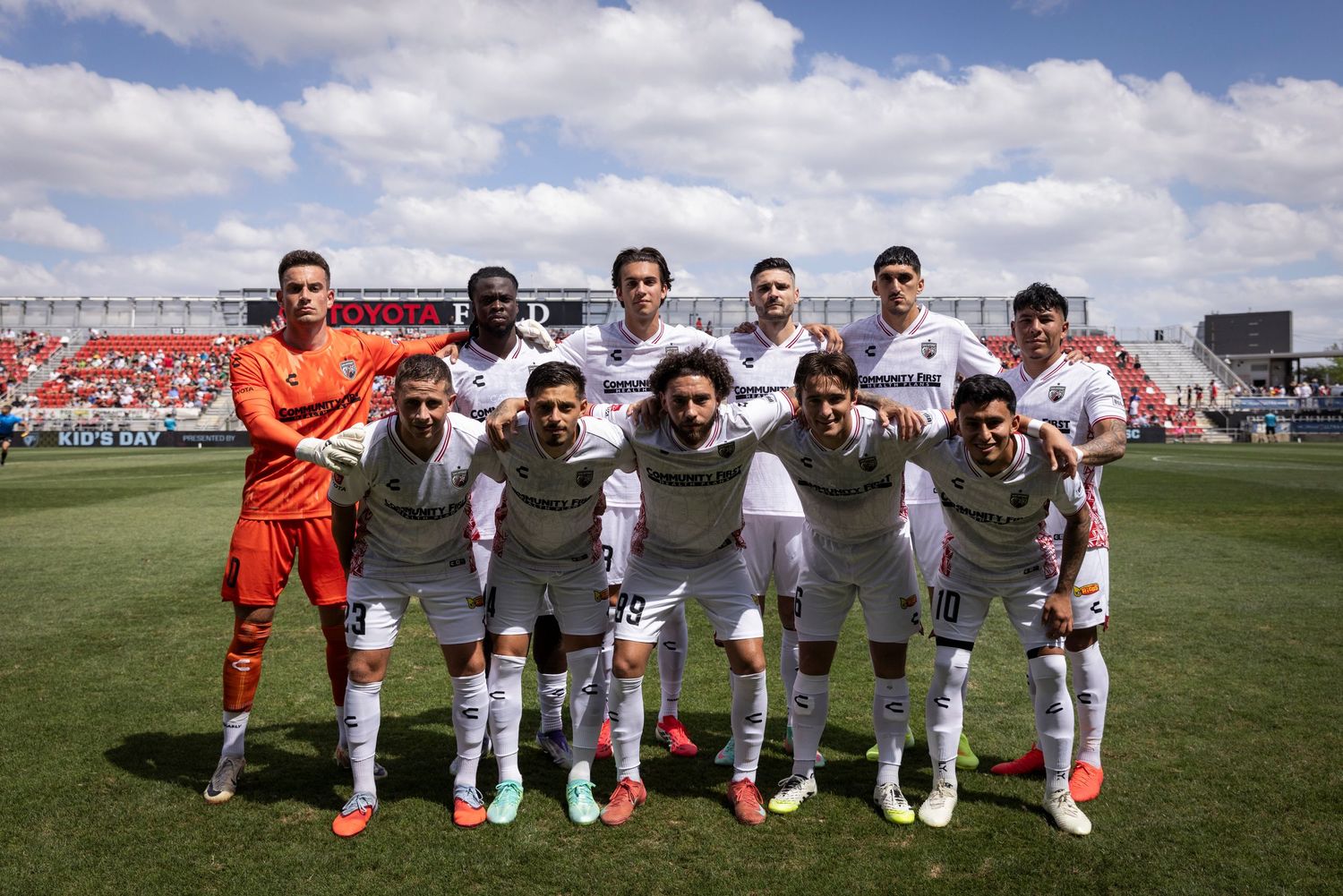 Soccer team poses for official pre-match photo on field, players in white uniforms with goalkeeper in orange jersey.