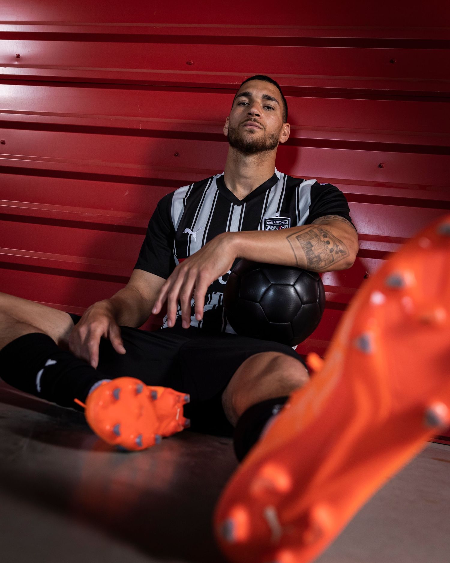 Soccer player rests with black ball and orange cleats against red metal wall.