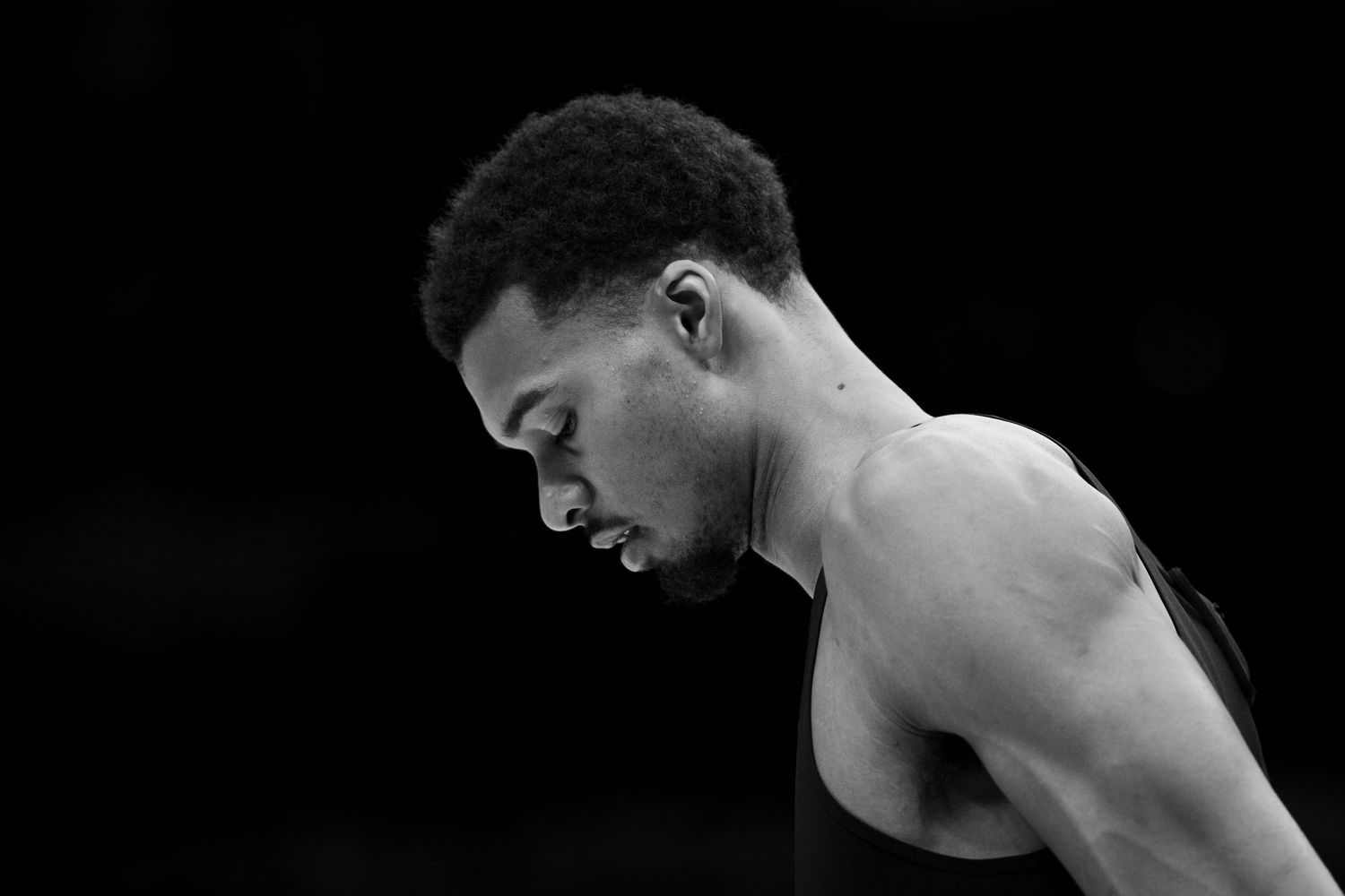 Black and white profile portrait of a shirtless athlete with short curly hair looking downward against a dark background.