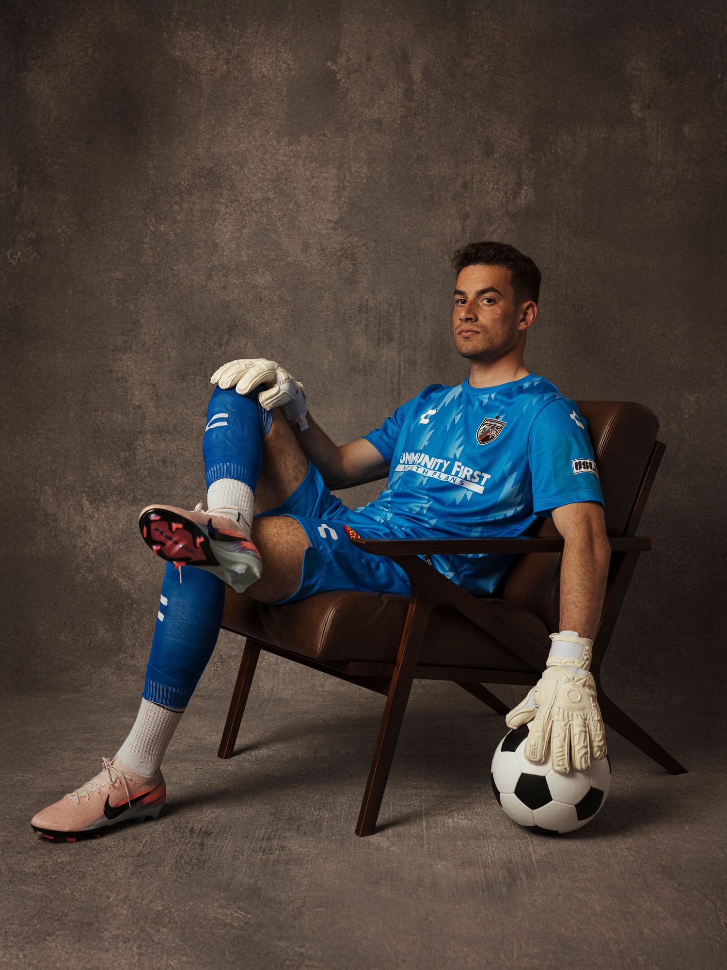 Soccer goalkeeper in blue uniform sitting in chair with ball and gloves, professional studio portrait photo.