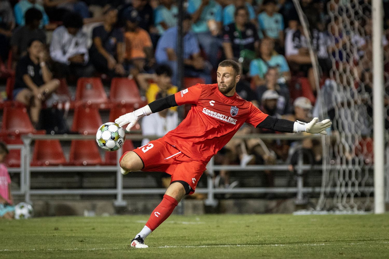 Soccer goalkeeper in red uniform kicking ball during a night match with fans in the background.