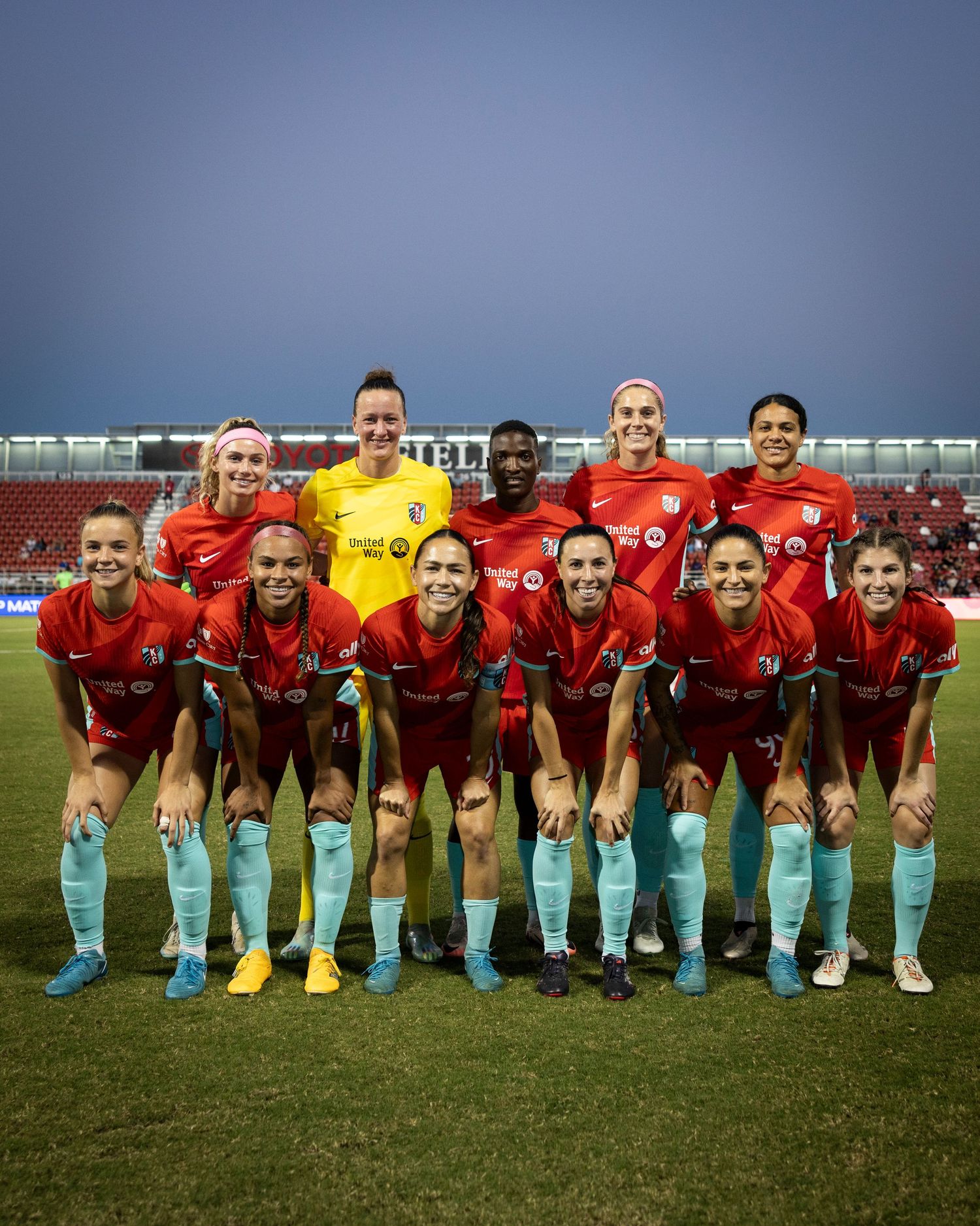 Soccer team wearing red jerseys and blue socks posing for group photo on field.