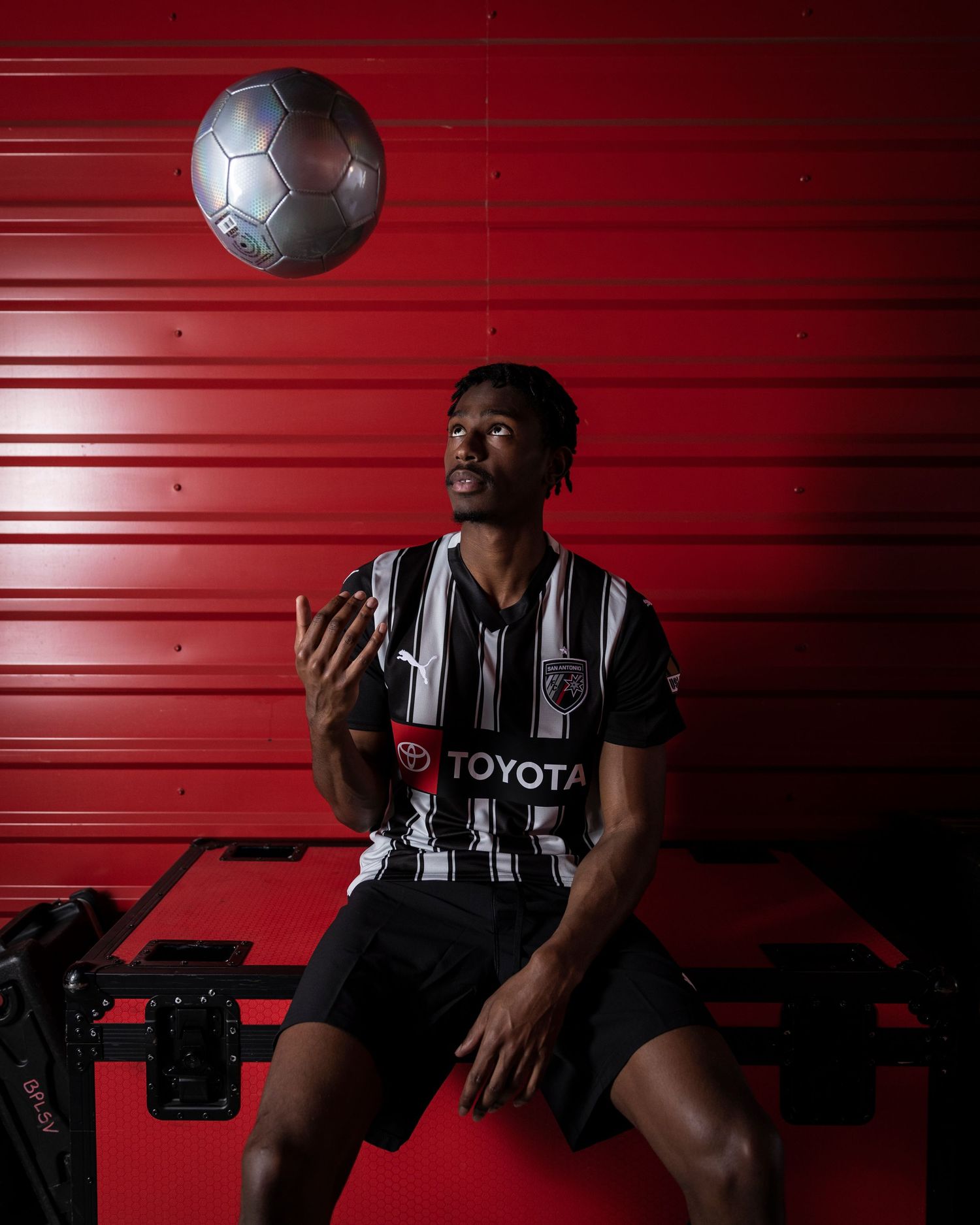 Soccer player in striped uniform juggles silver ball against red backdrop.