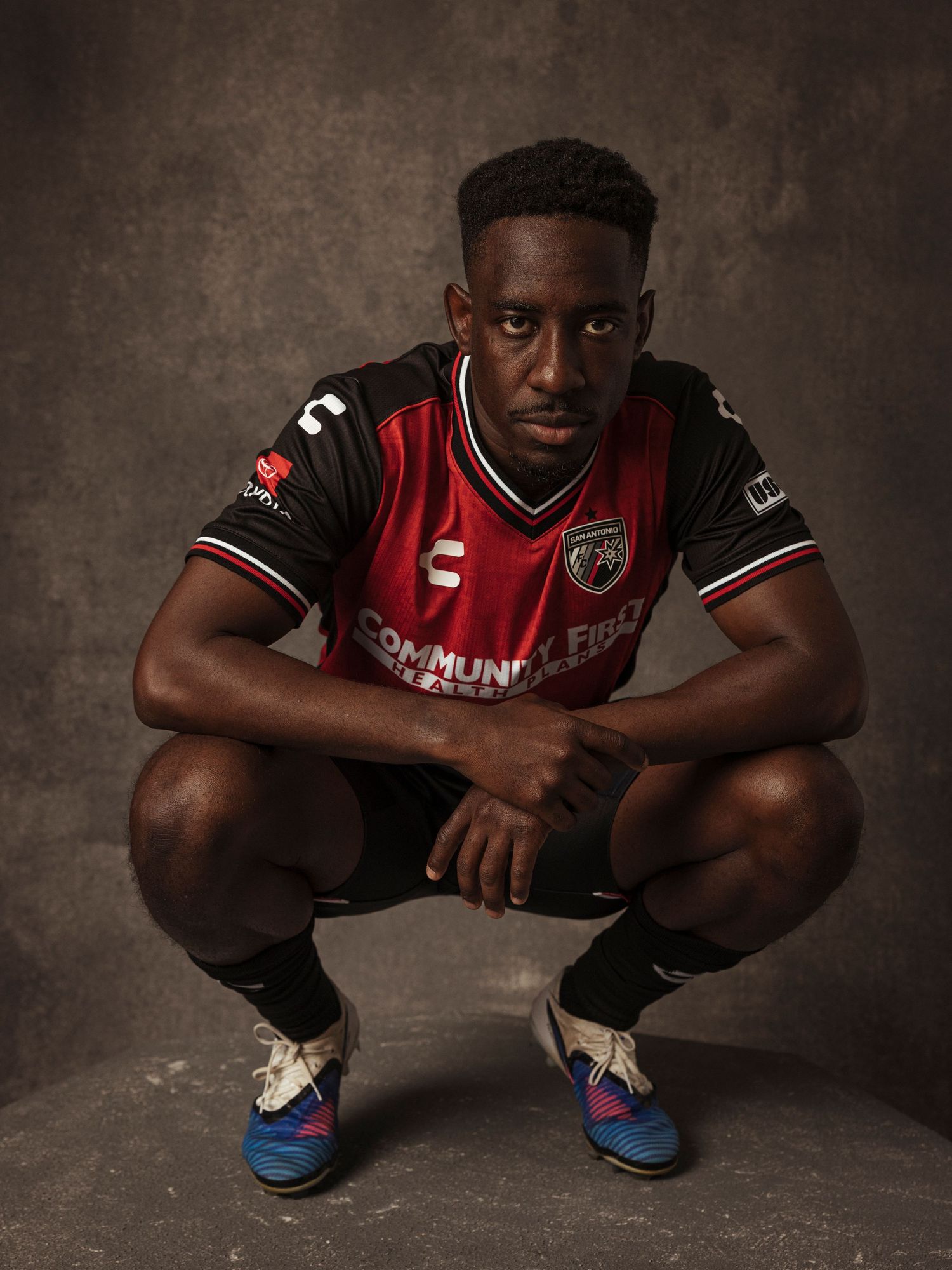 Soccer player in red and black Charly kit squatting confidently in a dramatic studio portrait pose.