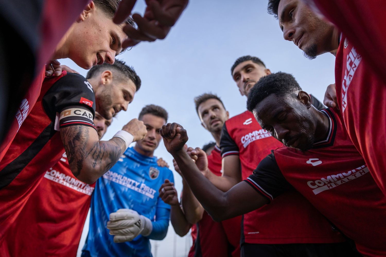 Soccer team huddles together in pre-match motivational circle on sunny day.