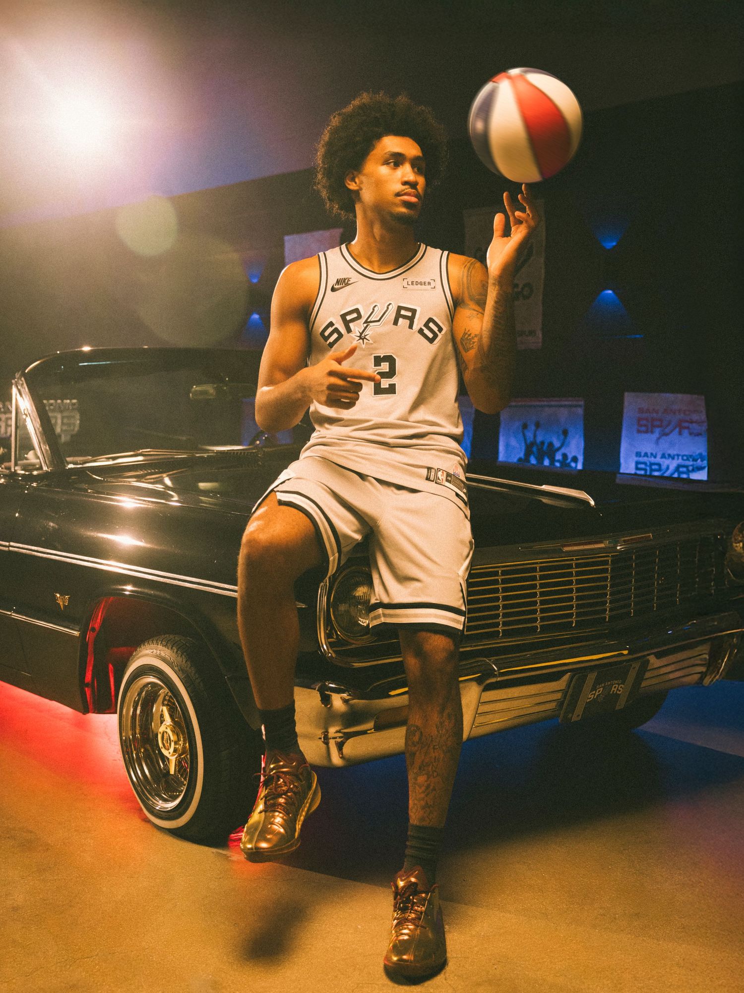 San Antonio Spurs player #2 spins a basketball on finger, posing in white uniform beside a classic black car.