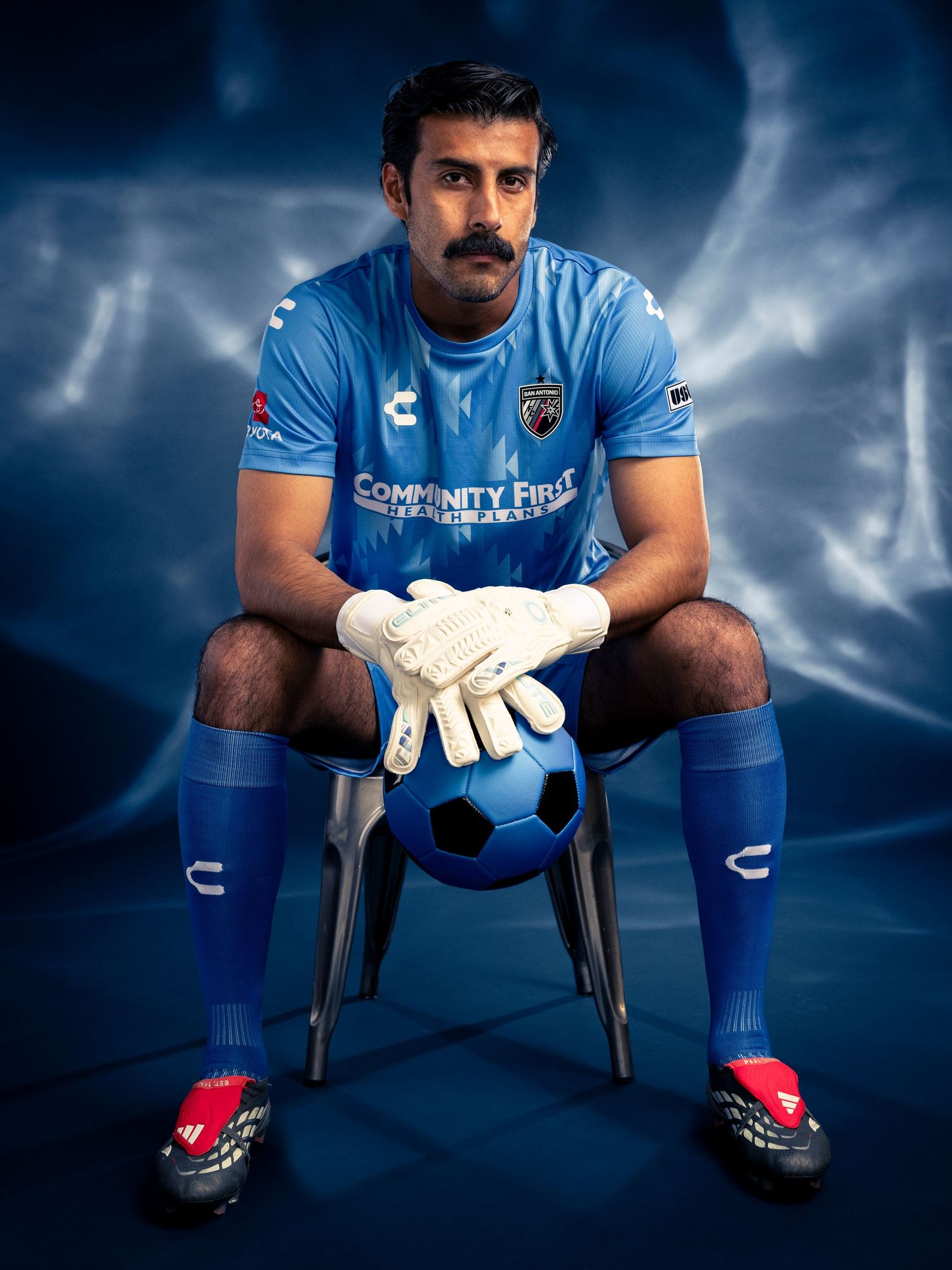 Soccer goalkeeper in blue Community First kit, wearing gloves, sitting with a ball in a dramatic blue studio setting.