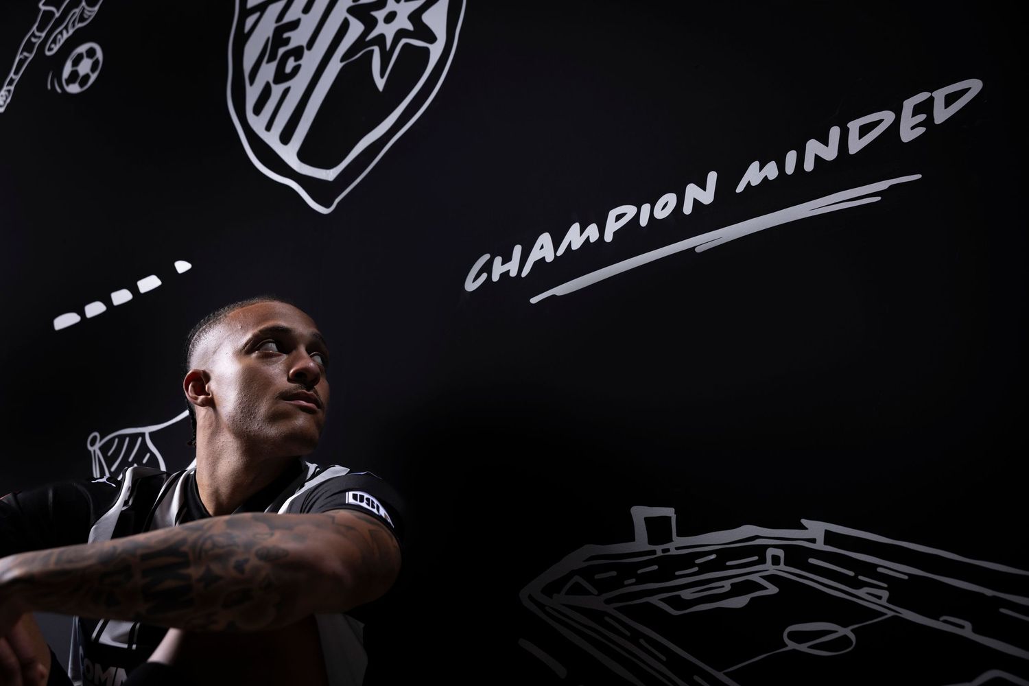 Soccer player in dark uniform poses against wall with champion minded text and drawings.