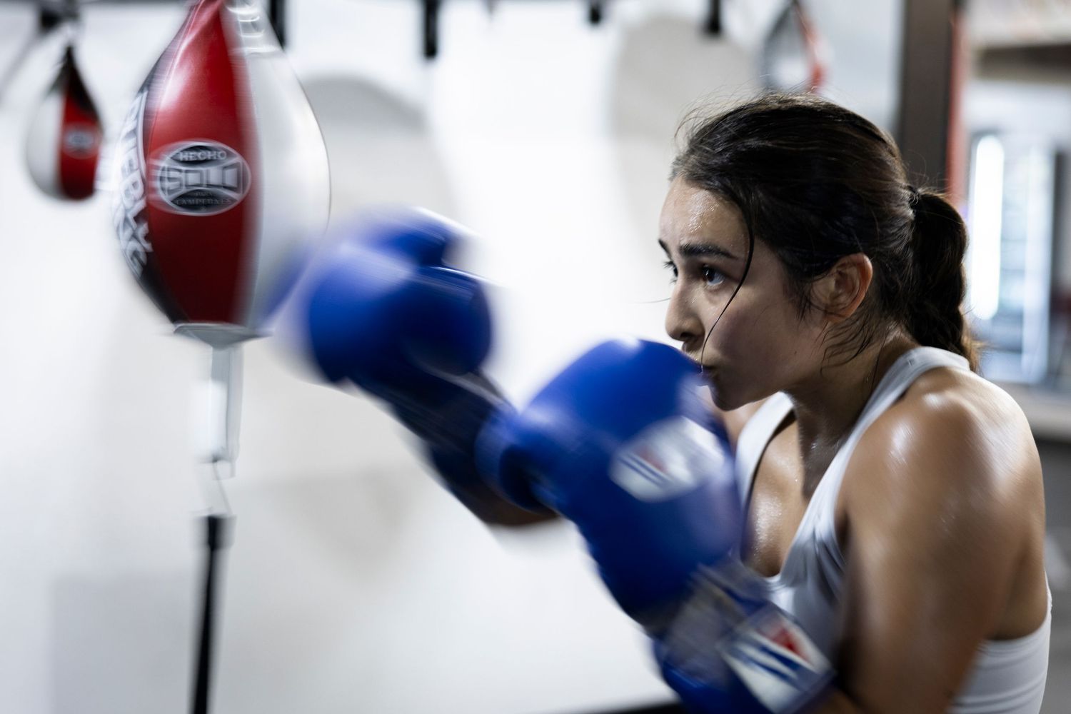 Boxer training with punching bag in a gym wearing blue gloves and white tank top.
