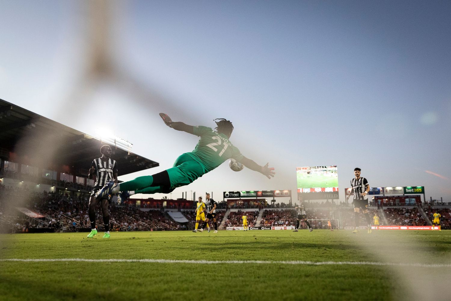 A dramatic soccer match moment captured through stadium netting during sunset.