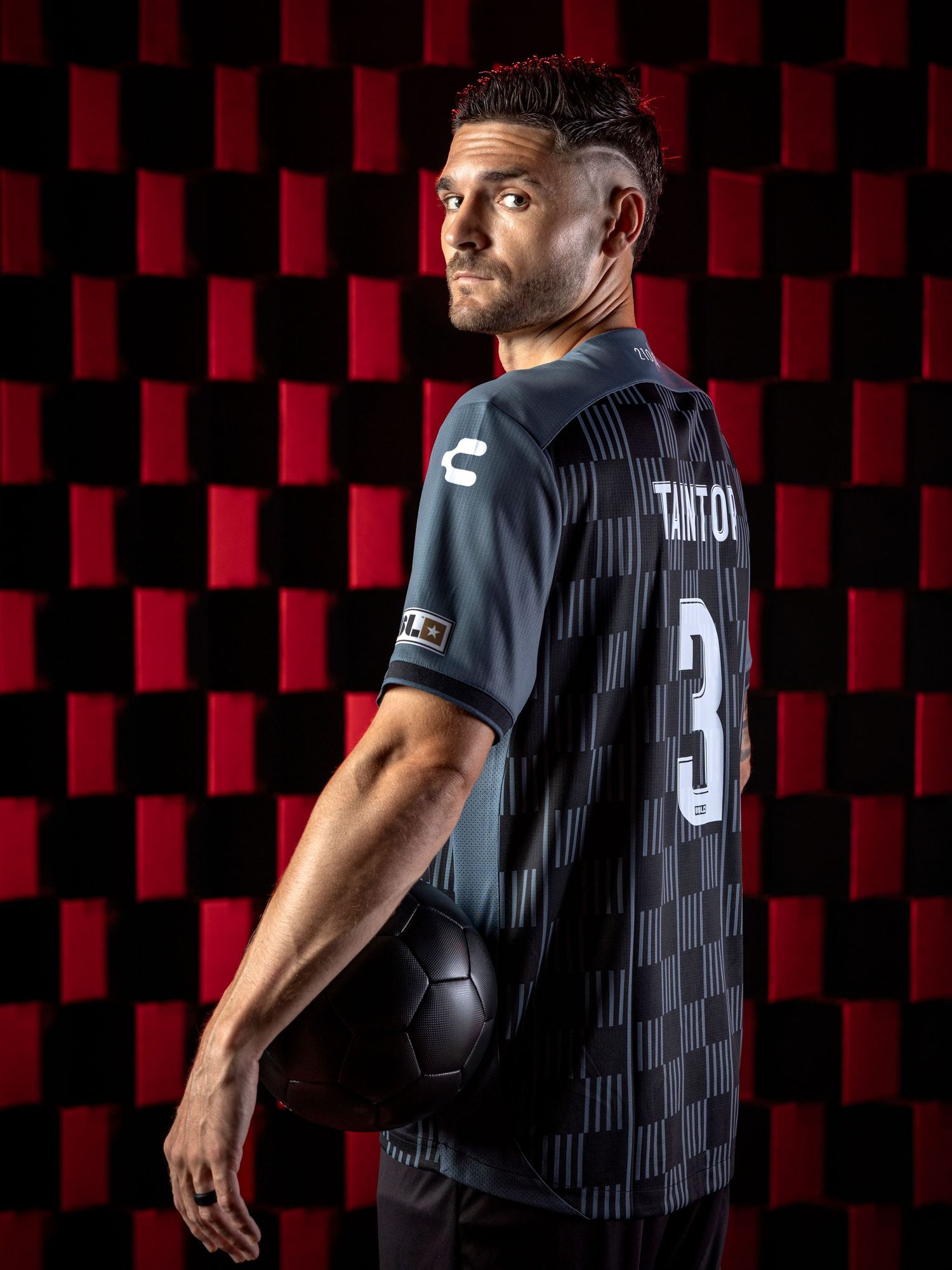 Soccer player in black jersey with number 3 poses against red and black checkered background holding a ball.