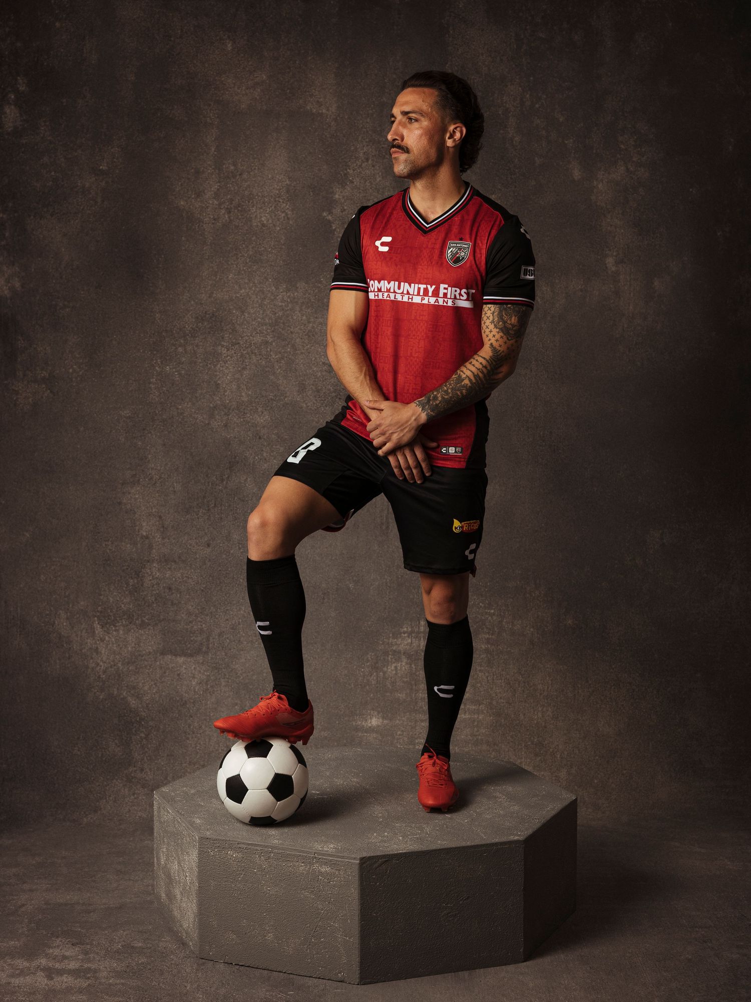 Soccer player in red and black jersey poses with ball on concrete platform in professional studio portrait.