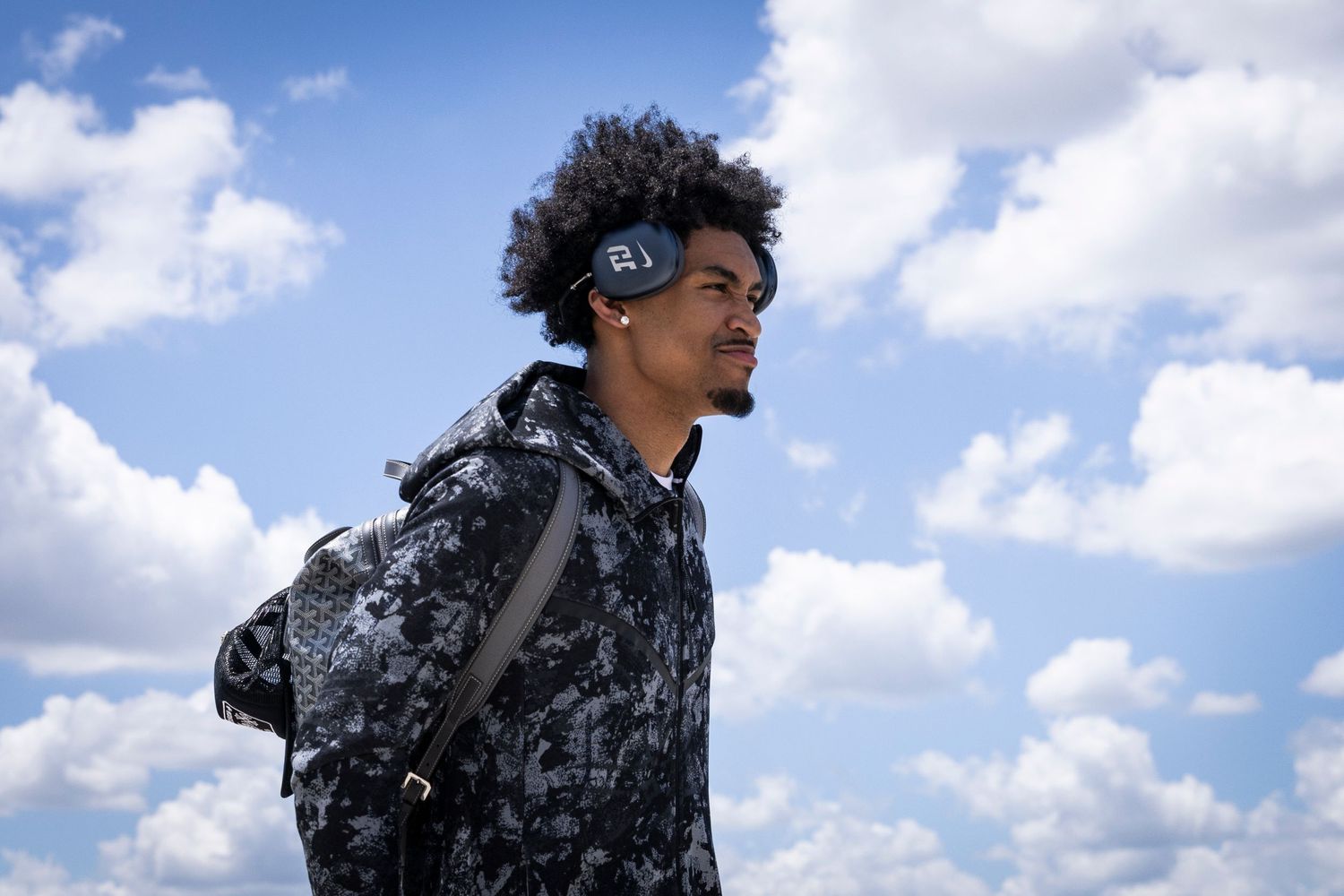 A person wearing a black and white patterned jacket stands against a bright blue sky with white clouds.