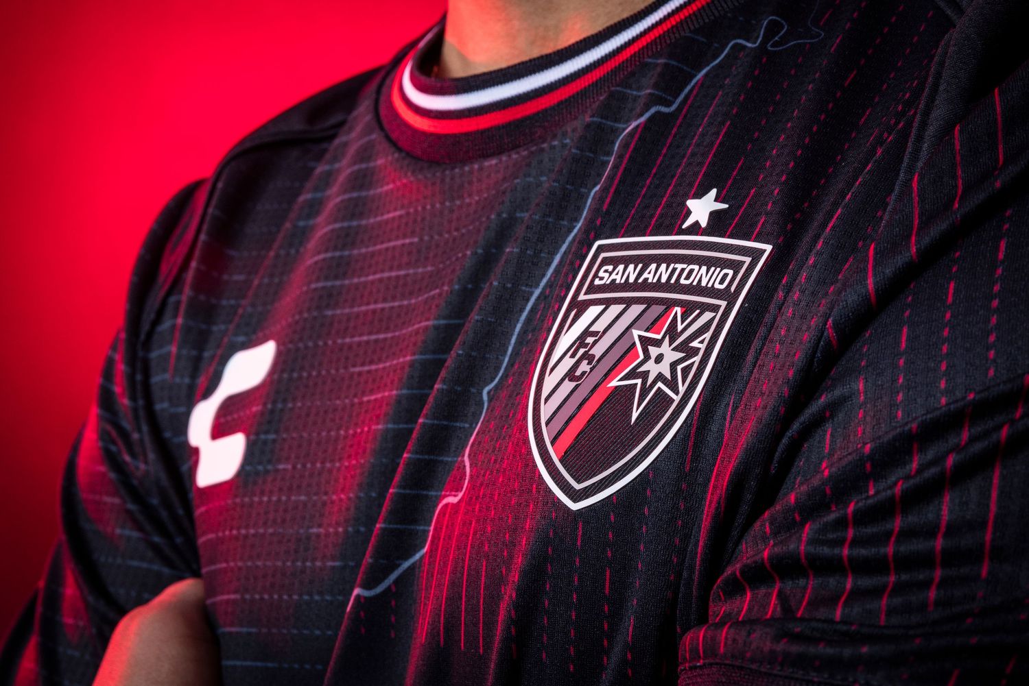 Close-up of black soccer jersey with red accents and team crest against red background.