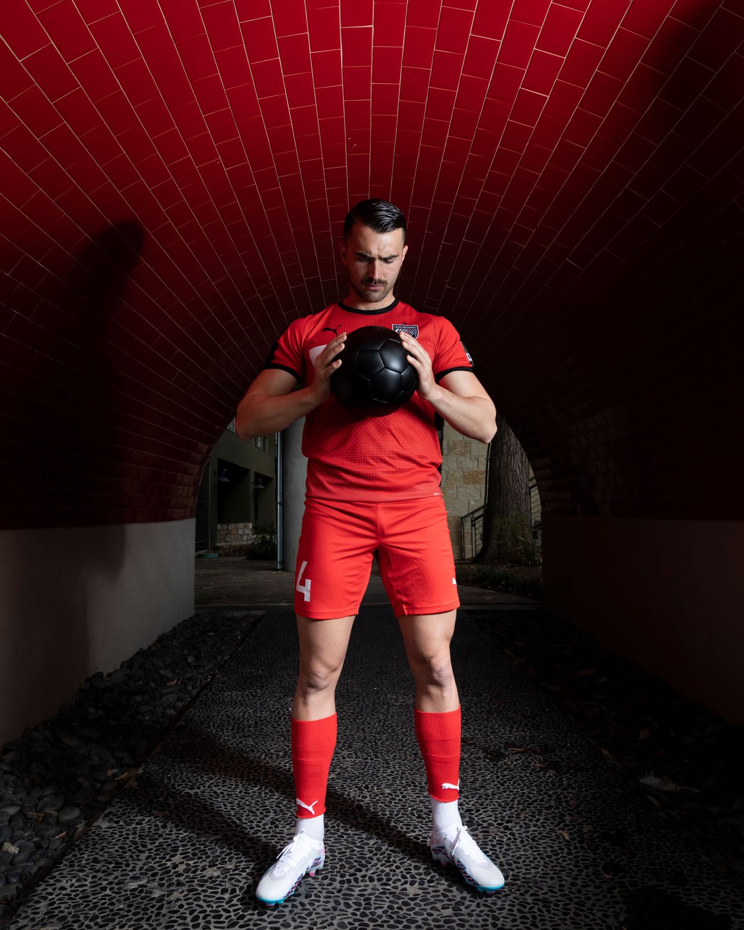 Soccer player in red uniform holds ball in dark tunnel with arched brick ceiling.