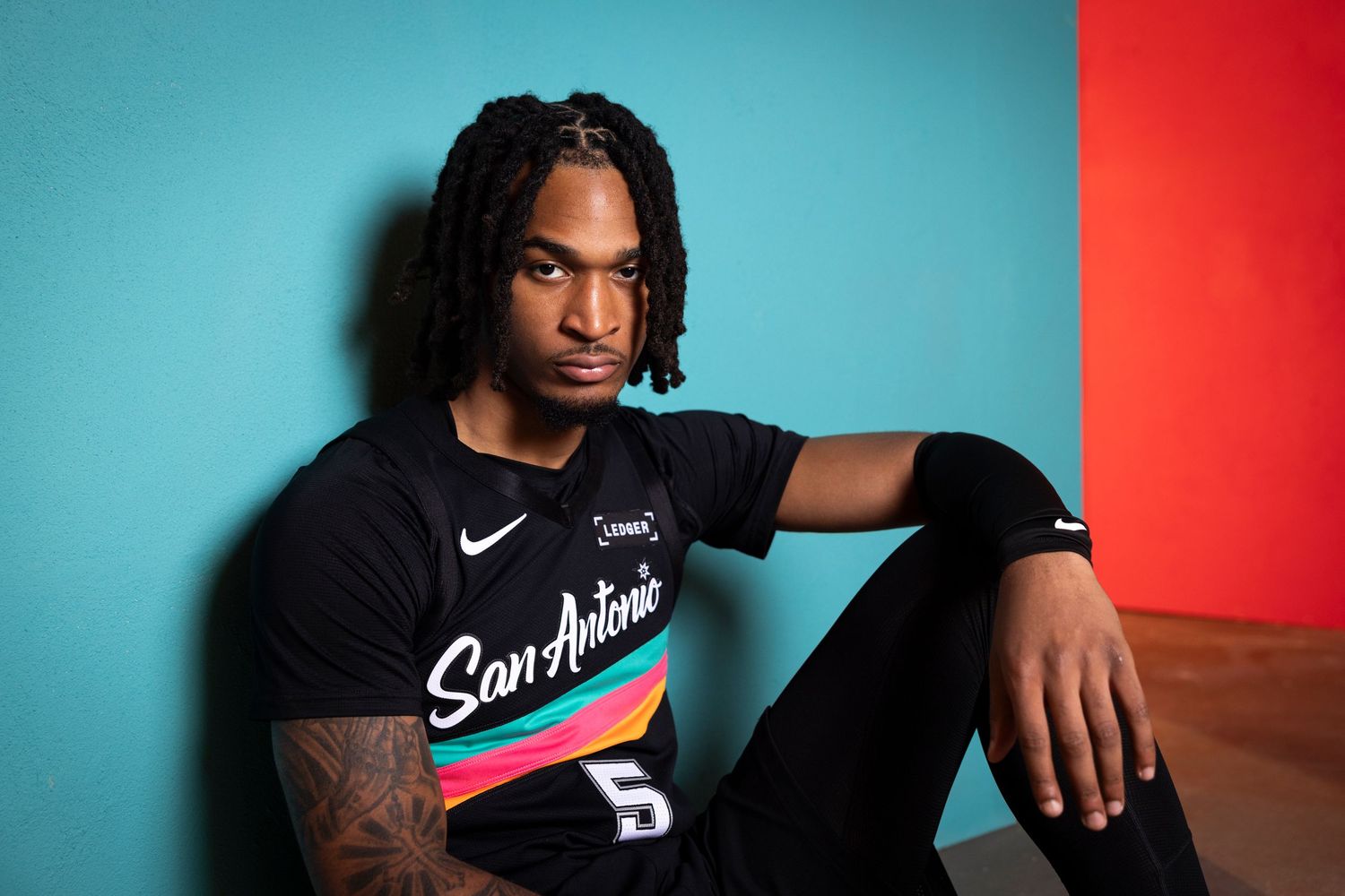 Basketball player wearing a black San Antonio jersey number 5, posing against a teal and red background.