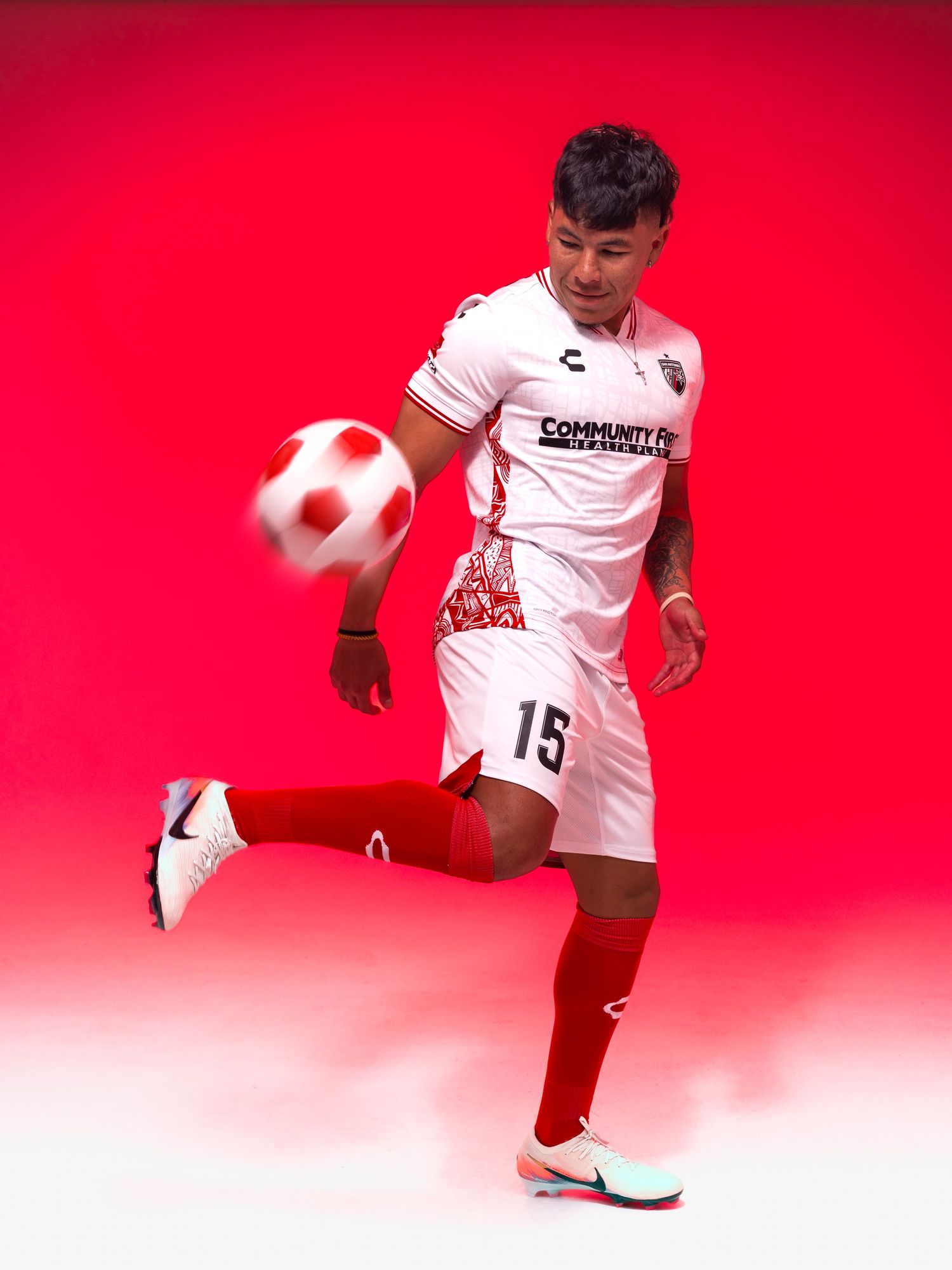 Soccer player in white #15 jersey and red socks skillfully juggling a ball against a vibrant red background.
