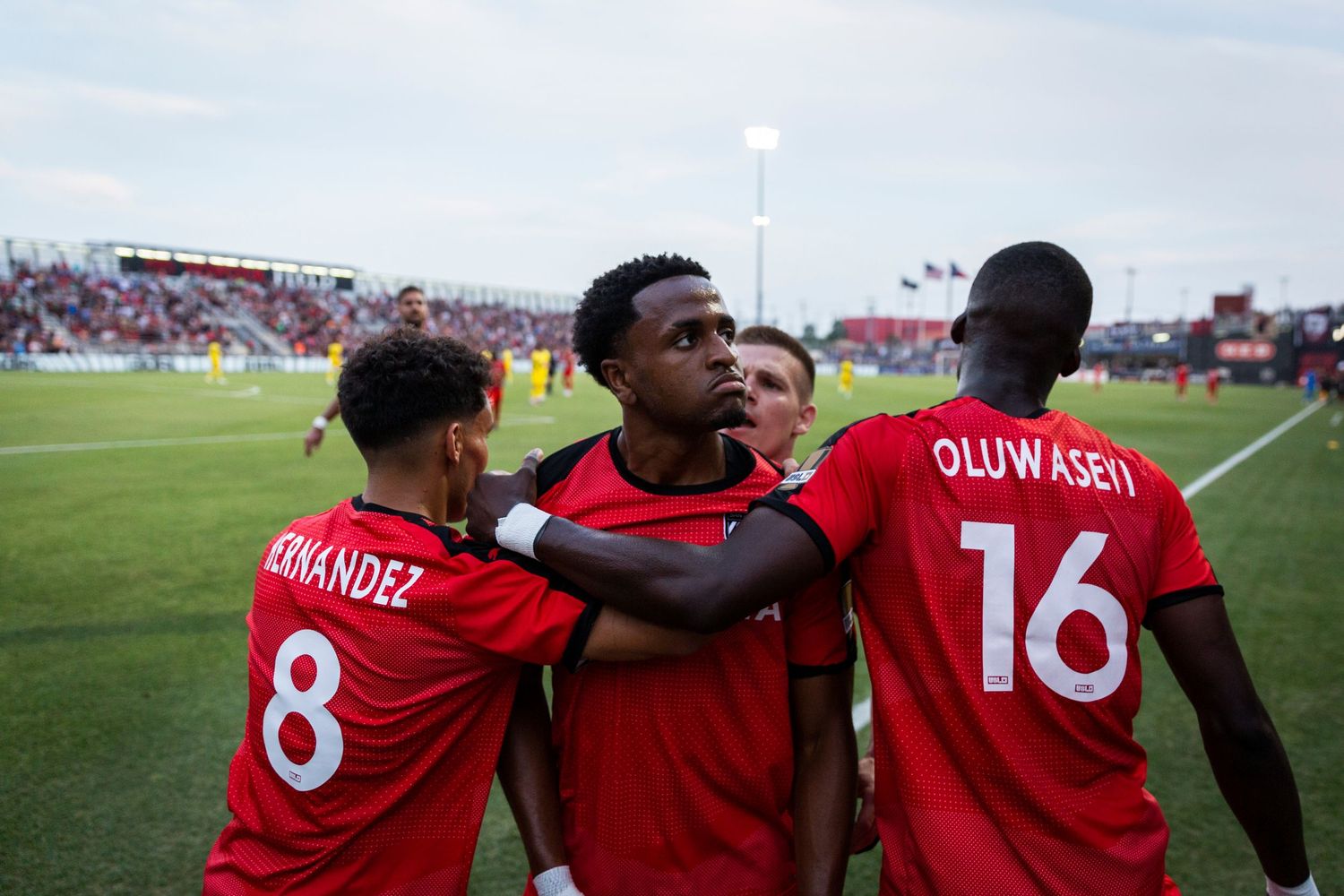 Three soccer teammates in red jerseys celebrate together on the field.