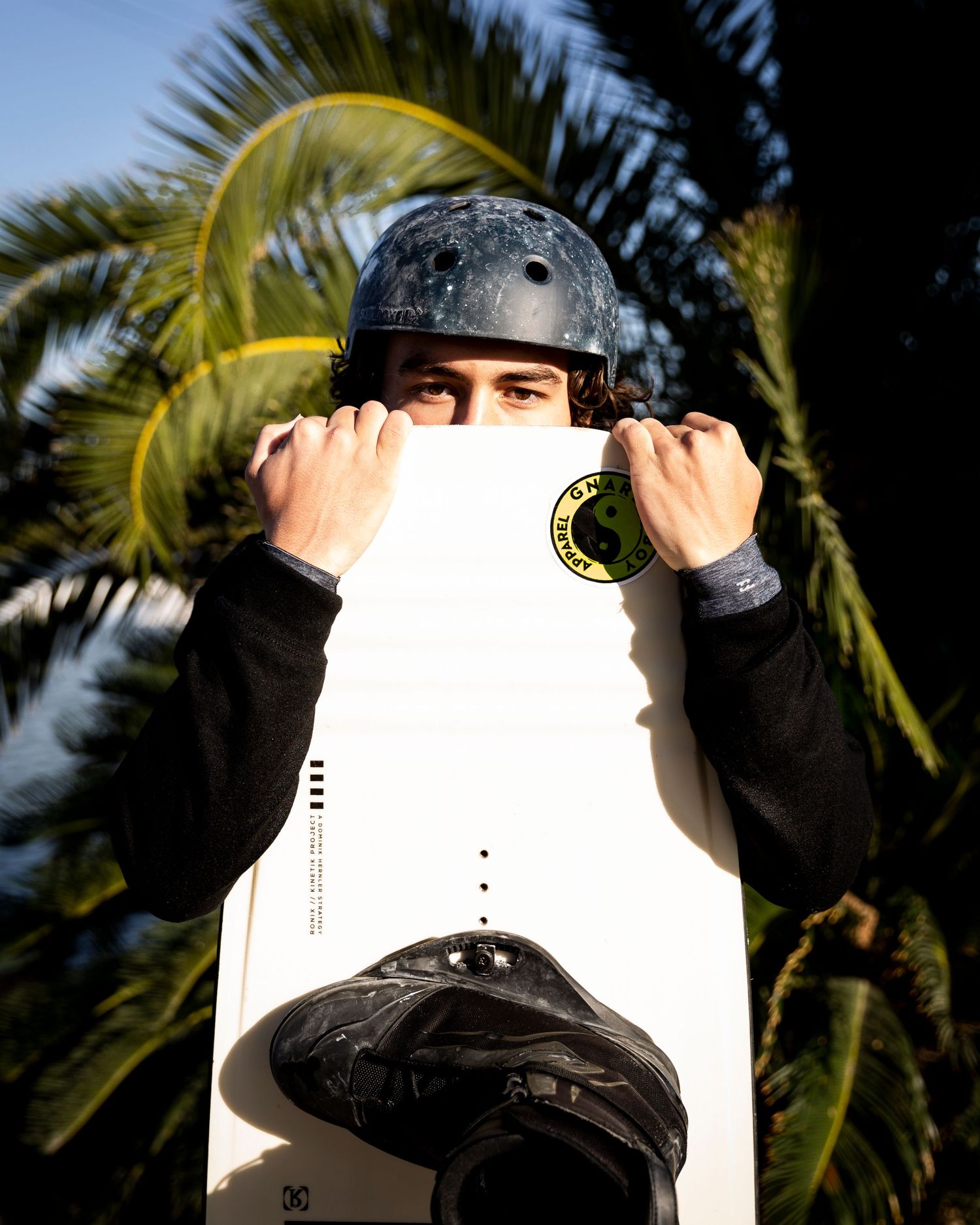 Skater in protective helmet holding a white skateboard deck in front of palm trees.
