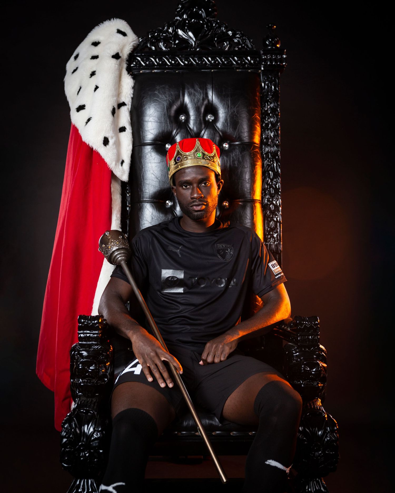 Soccer player poses regally in black throne wearing crown and red cape.