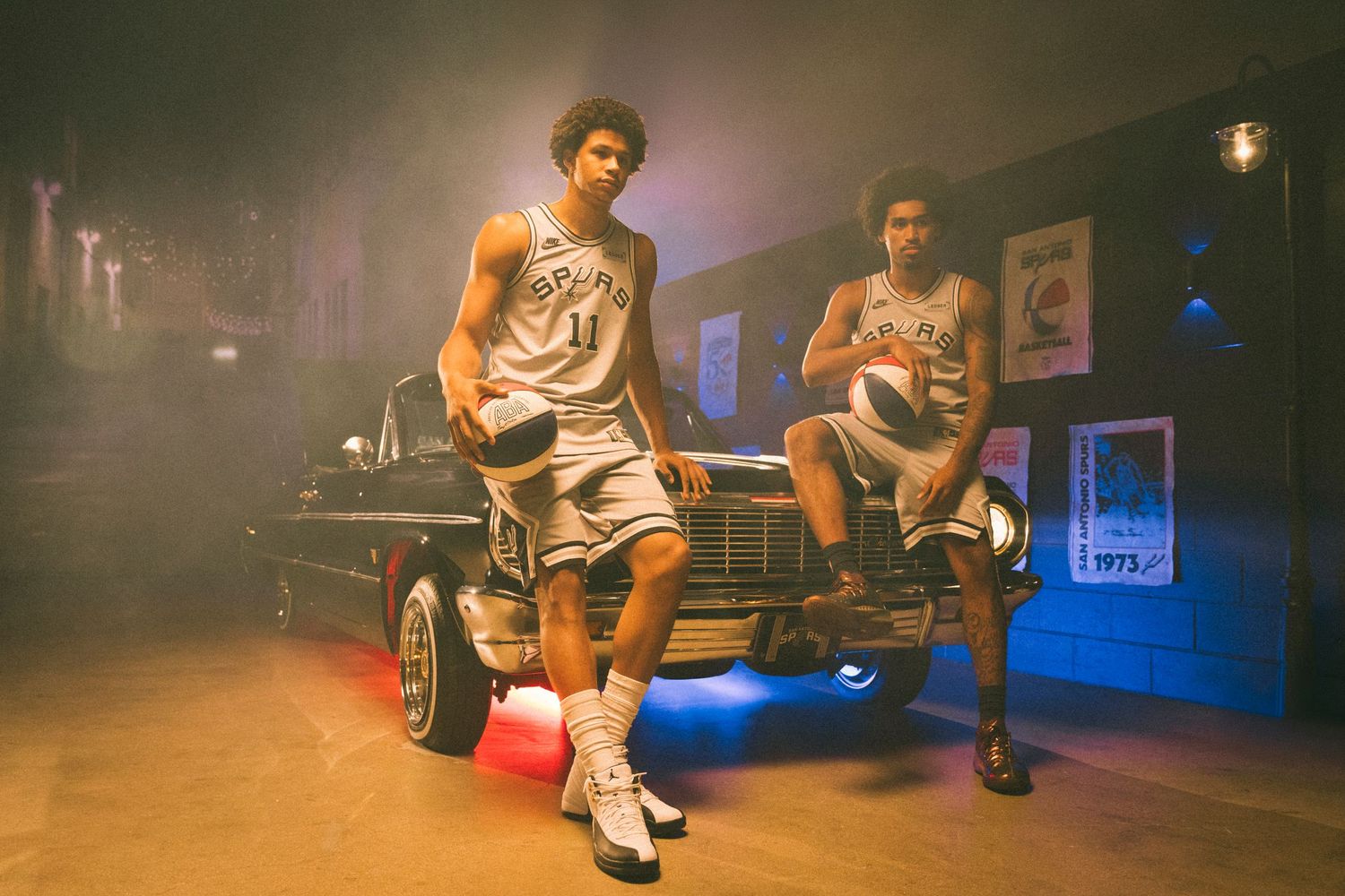 Two basketball players in San Antonio Spurs jerseys hold basketballs beside a classic lowrider car in a moody, neon-lit garage setting.