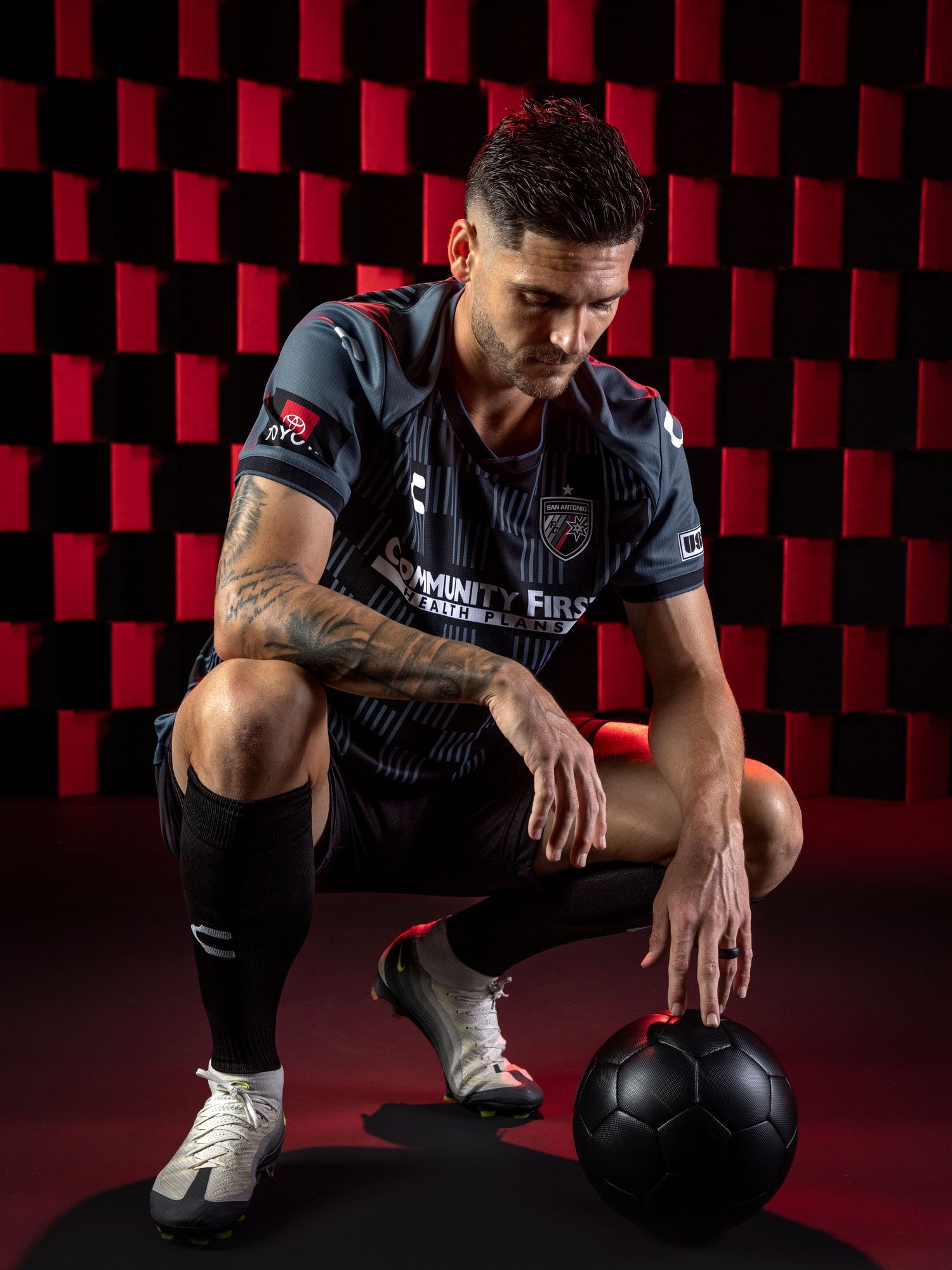 Soccer player in black uniform kneels with black ball against geometric red and black background pattern.