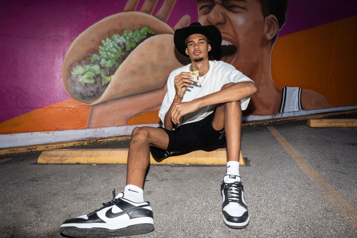 Casual portrait of person sitting against colorful mural wall wearing black shorts and Nike sneakers.