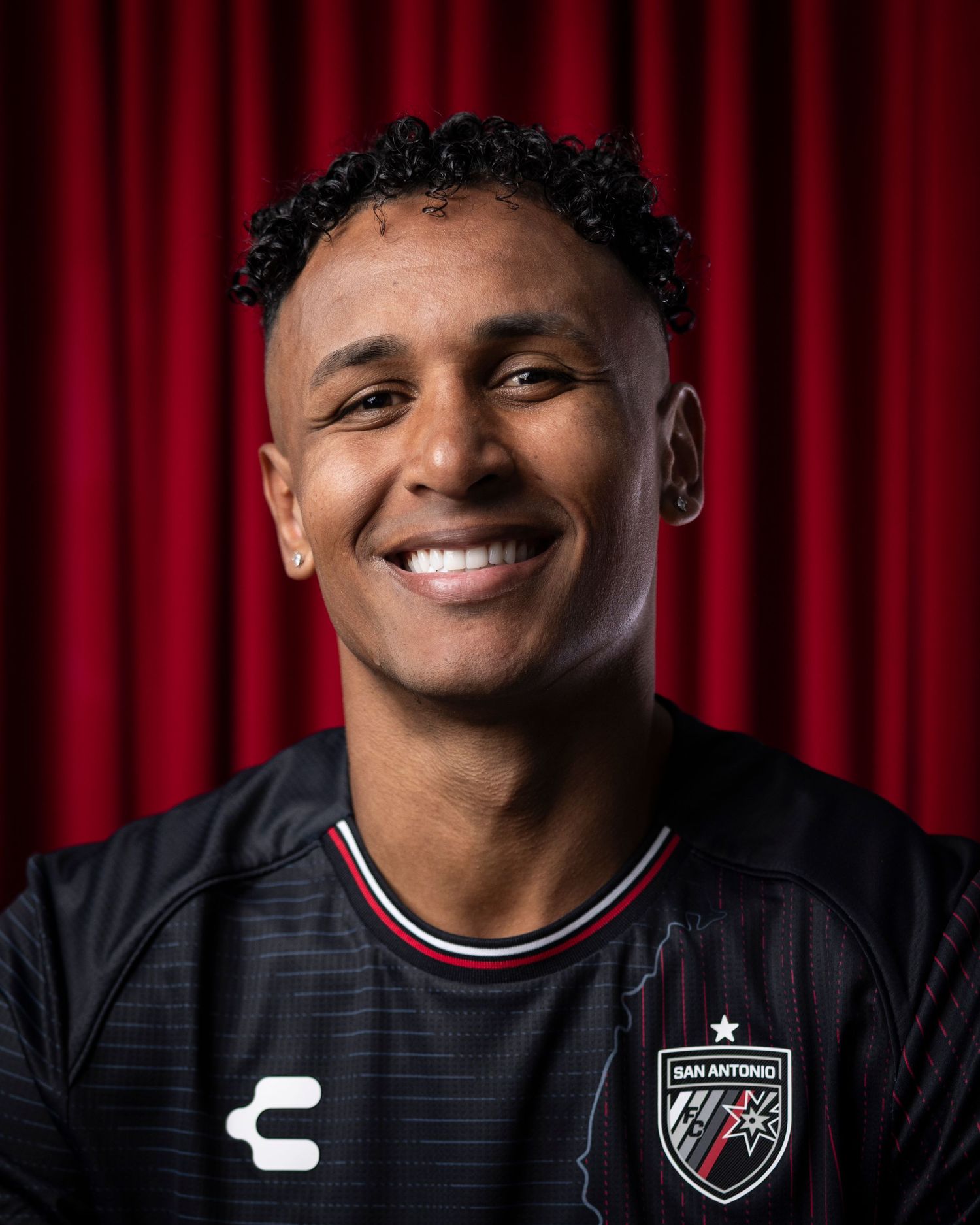Professional soccer player smiles warmly in team portrait against red backdrop.