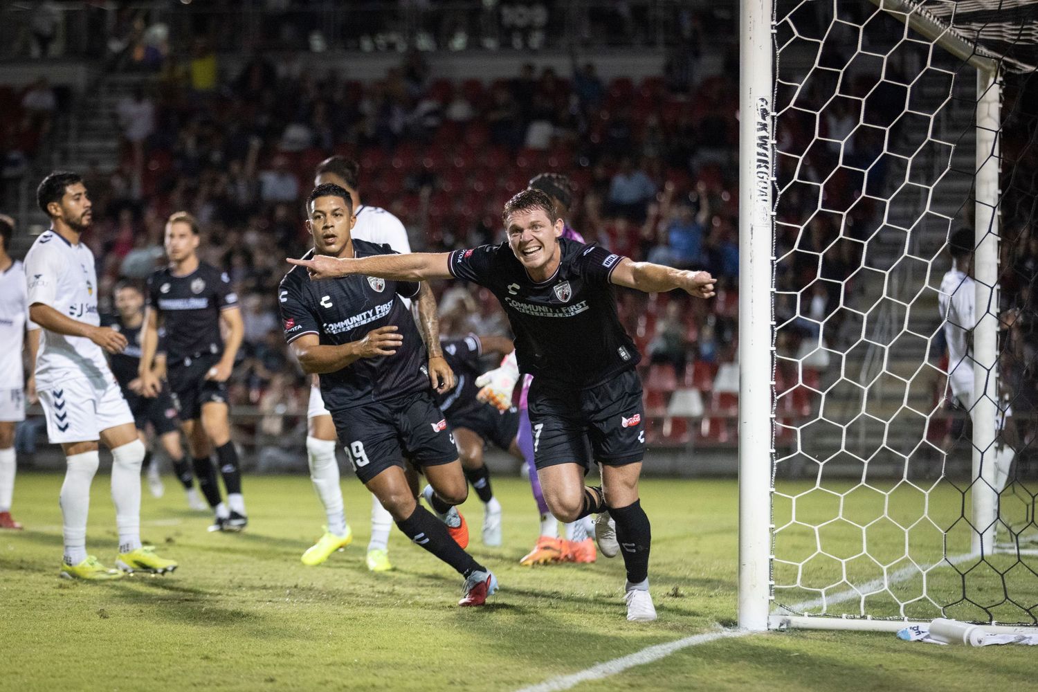 Two soccer players celebrating a goal near the net during a competitive night match.