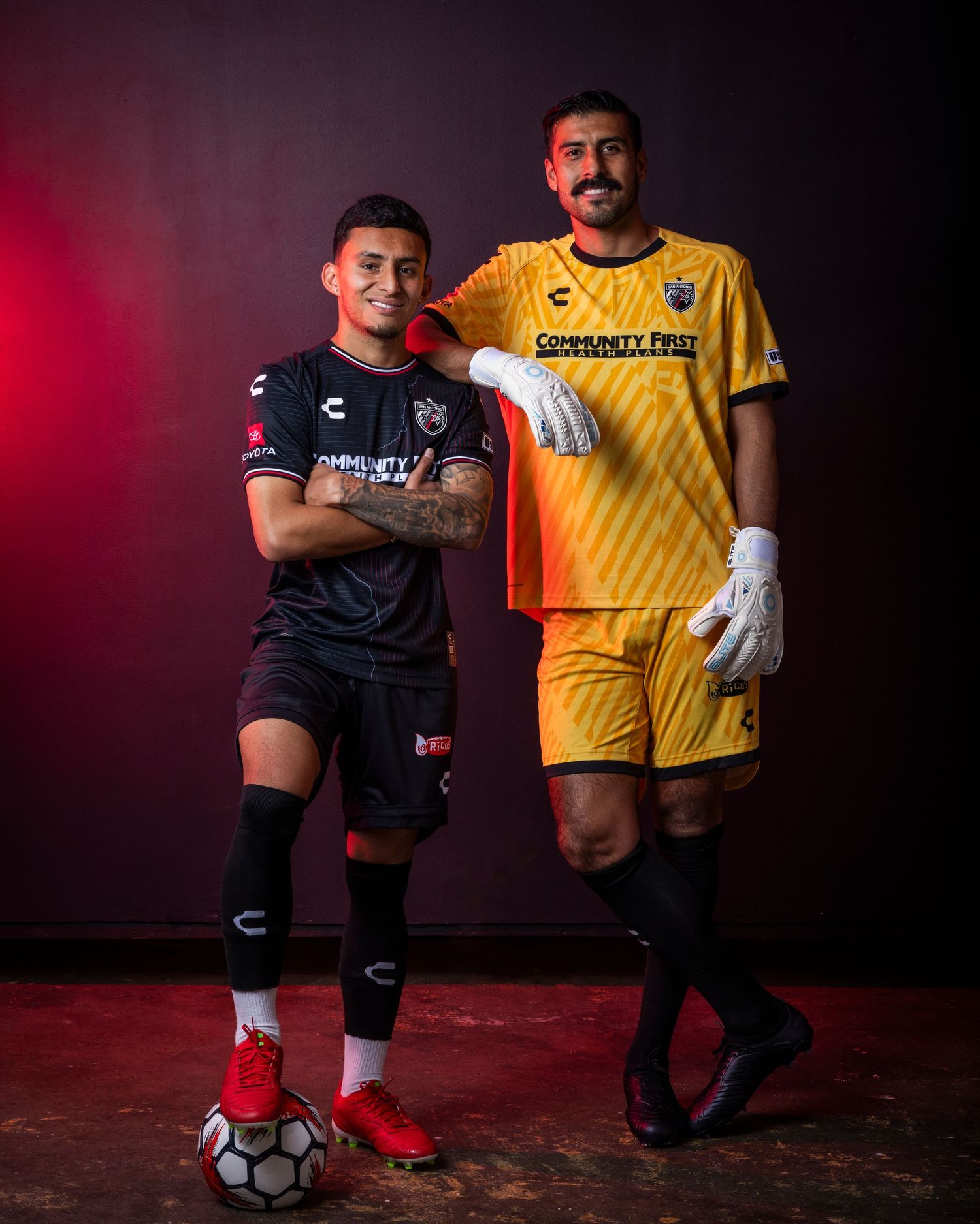 Two soccer players pose together wearing contrasting black and yellow team uniforms against a red backdrop.