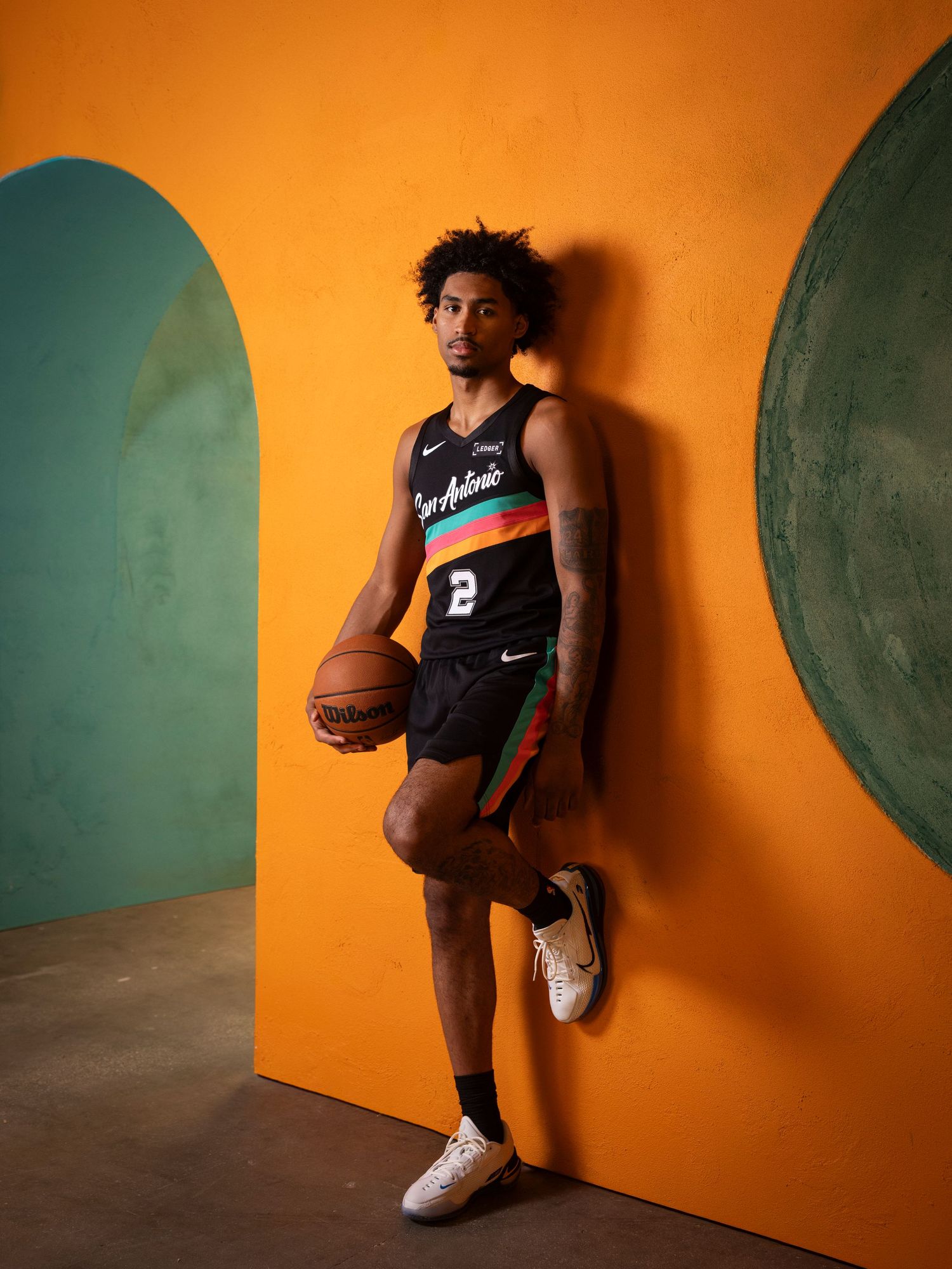 Male basketball player in San Antonio Nike jersey holding a ball against a vibrant orange wall.