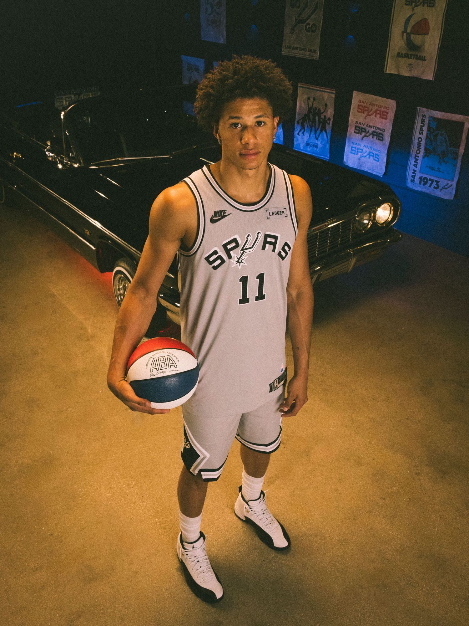 San Antonio Spurs player #11 poses in gray uniform holding a basketball in front of a vintage car.