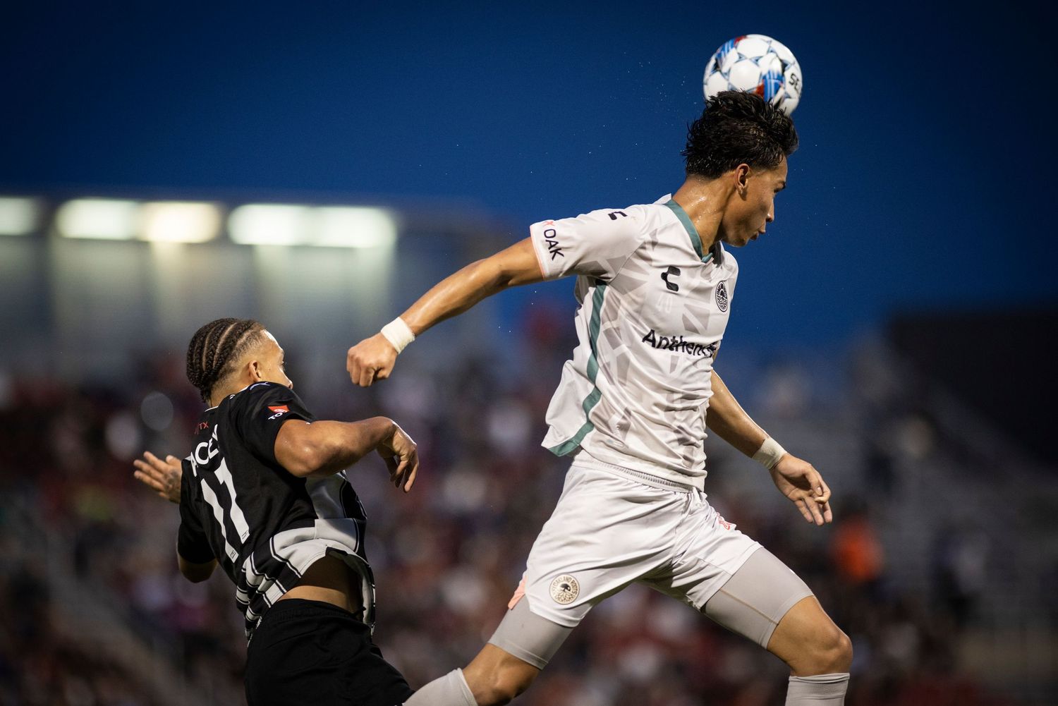 Two soccer players jump high to compete for a header during a night match.
