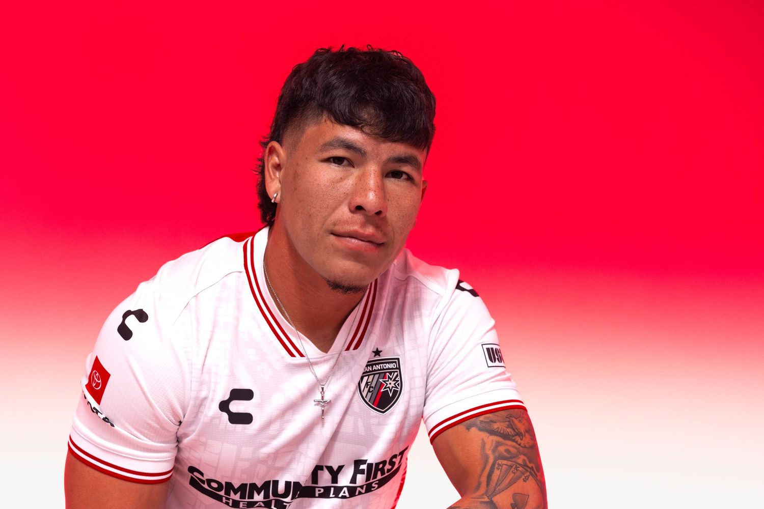 Soccer player in white Charly kit poses against red background, sporting tattoos and a confident expression.