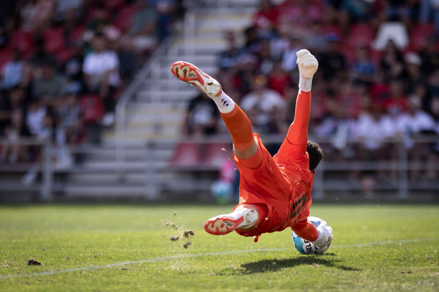 A goalkeeper in red dives to make a save during a sunny outdoor soccer match.