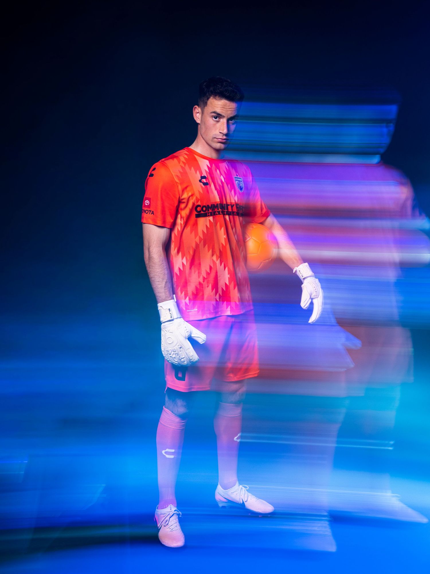 Goalkeeper poses in vibrant orange-pink jersey with colorful motion blur trails on dark background.