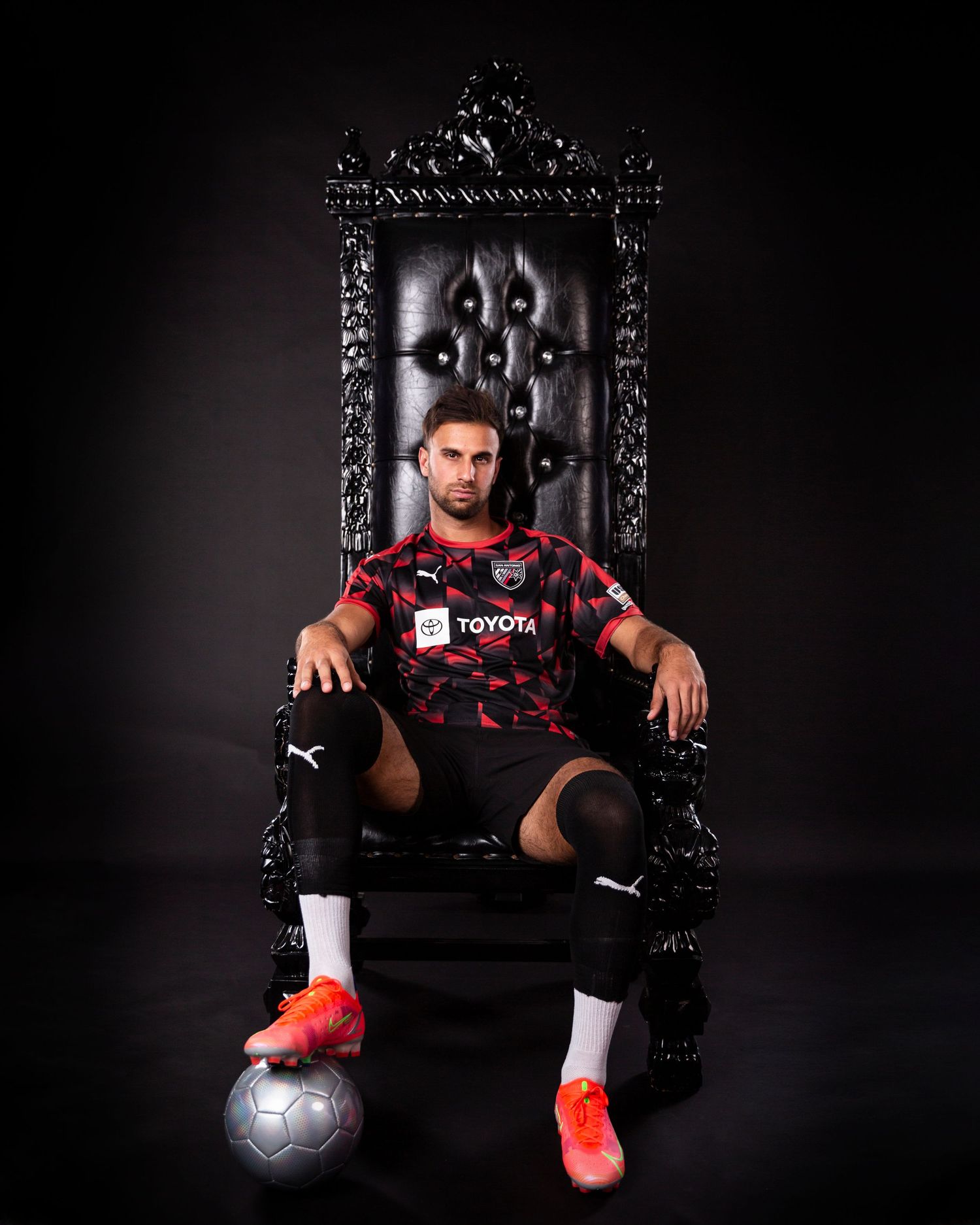 Soccer player sits on ornate throne with ball at feet wearing red and black uniform.