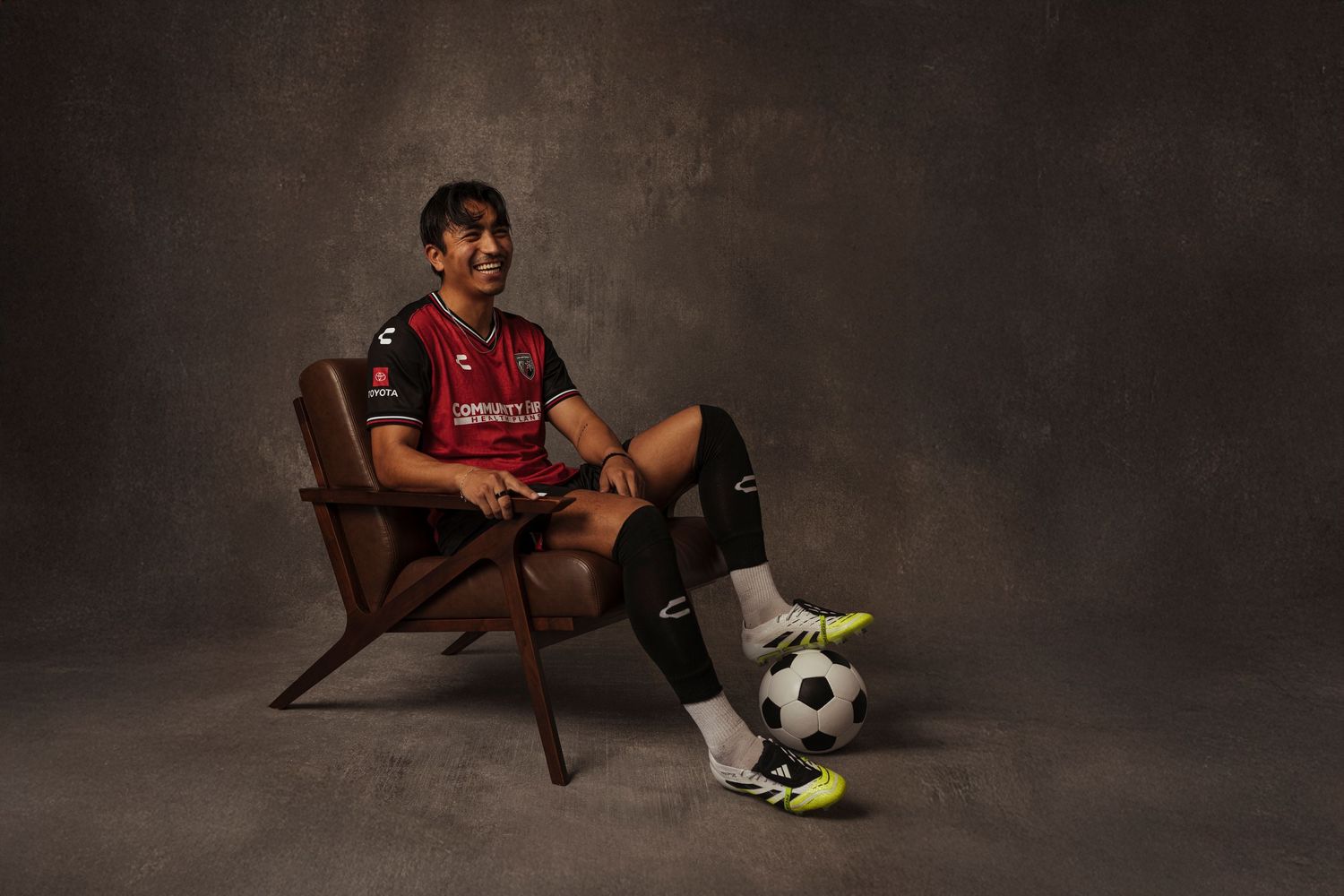 Soccer player in red jersey sits in wooden chair with ball at feet in professional studio portrait.