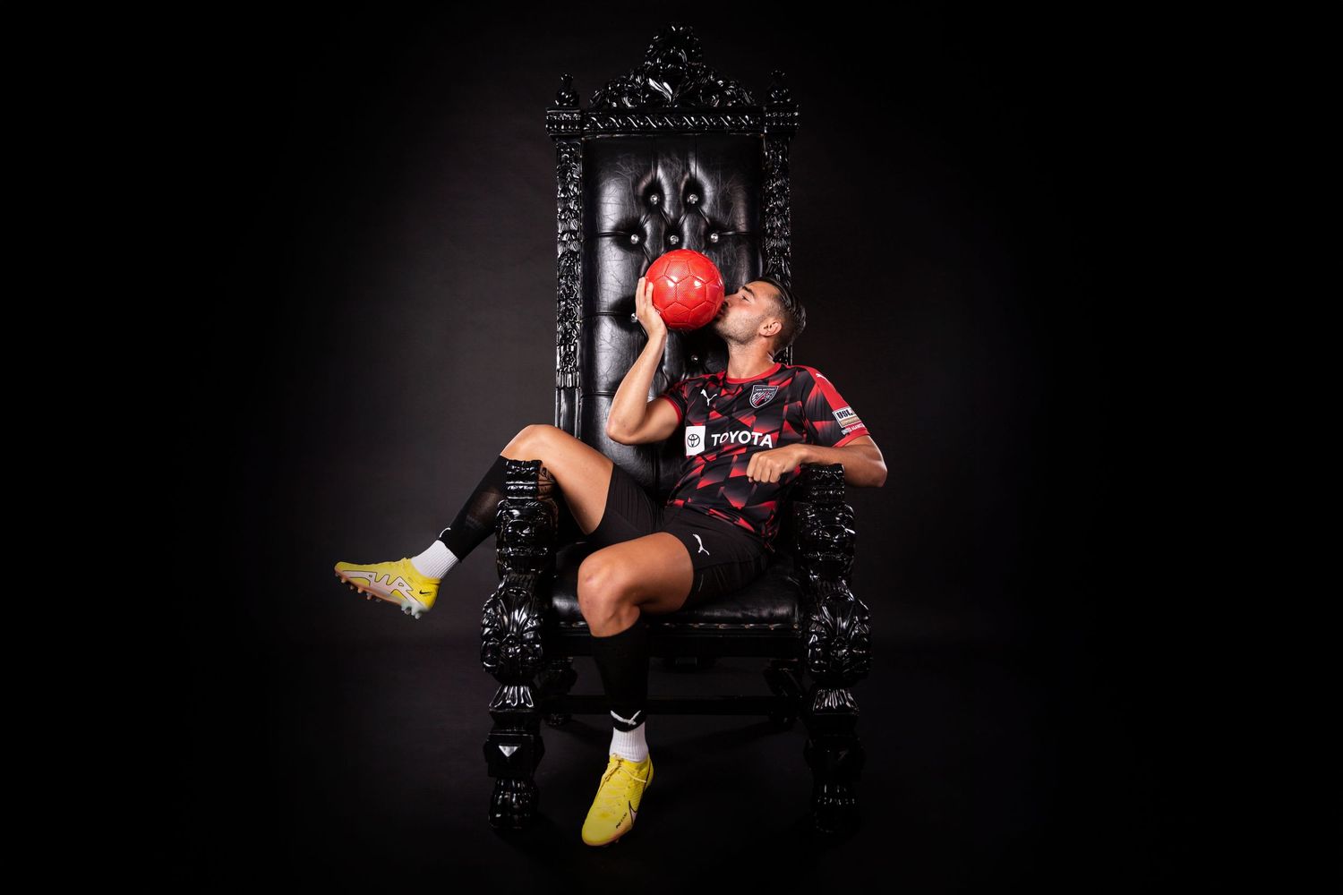 Soccer player seated on ornate black throne holding red ball in dramatic portrait.