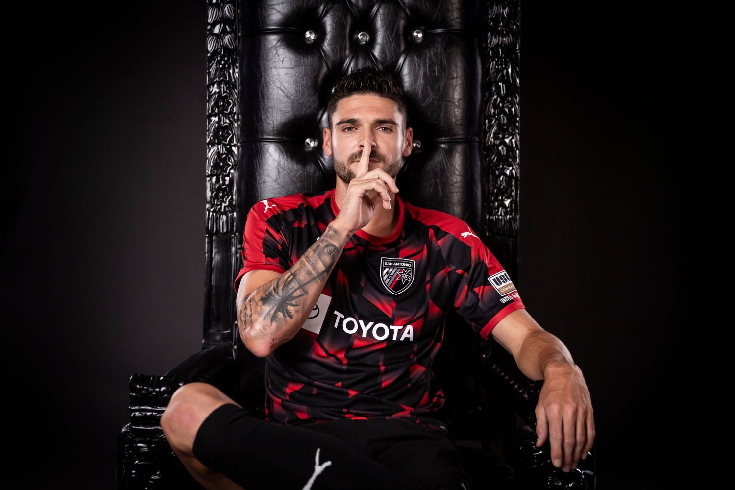 Soccer player poses in a dramatic black throne chair wearing a red and black Toyota jersey.