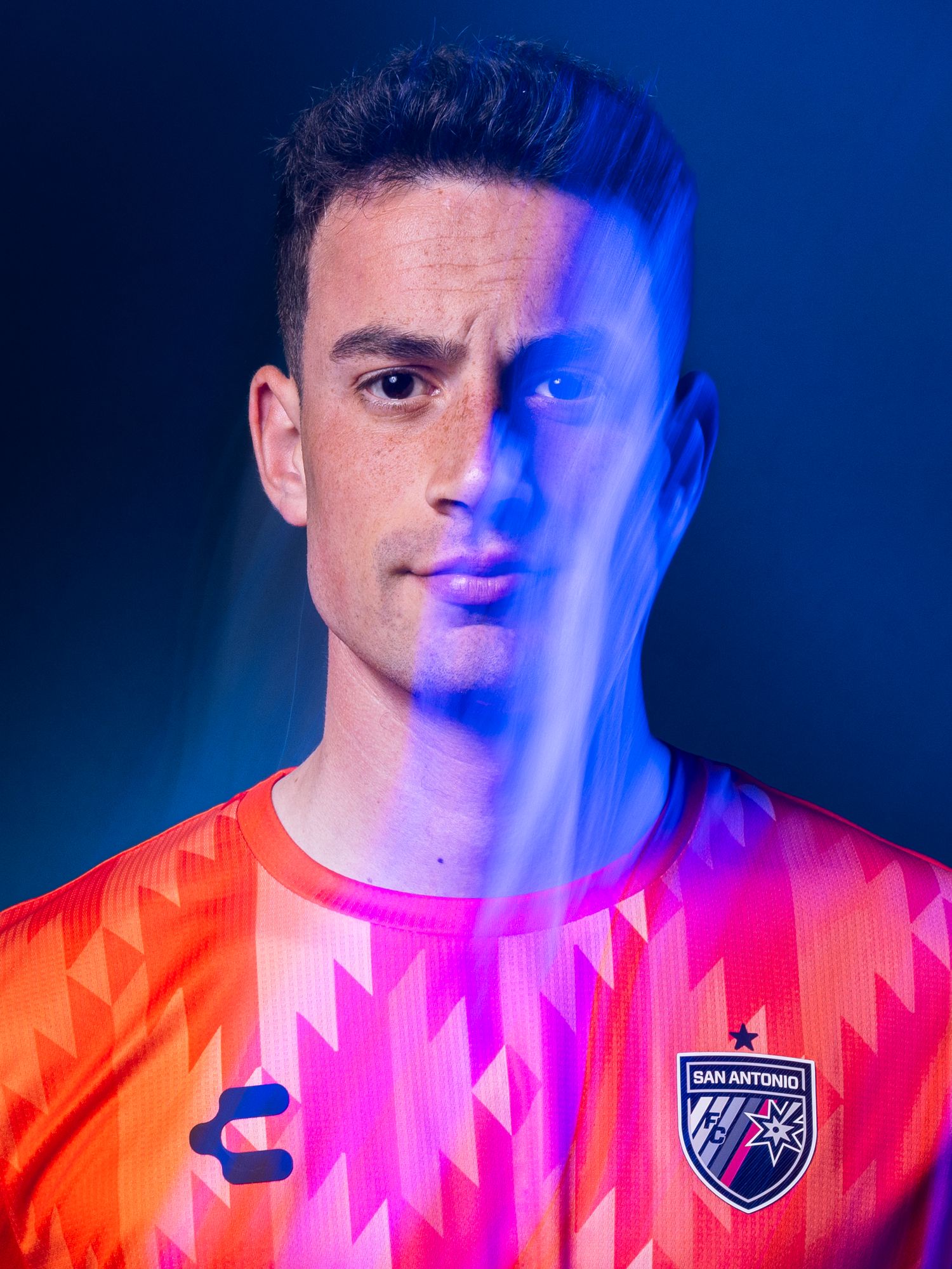 A male soccer player poses in a vibrant orange San Antonio FC jersey with geometric patterns under dramatic blue lighting.