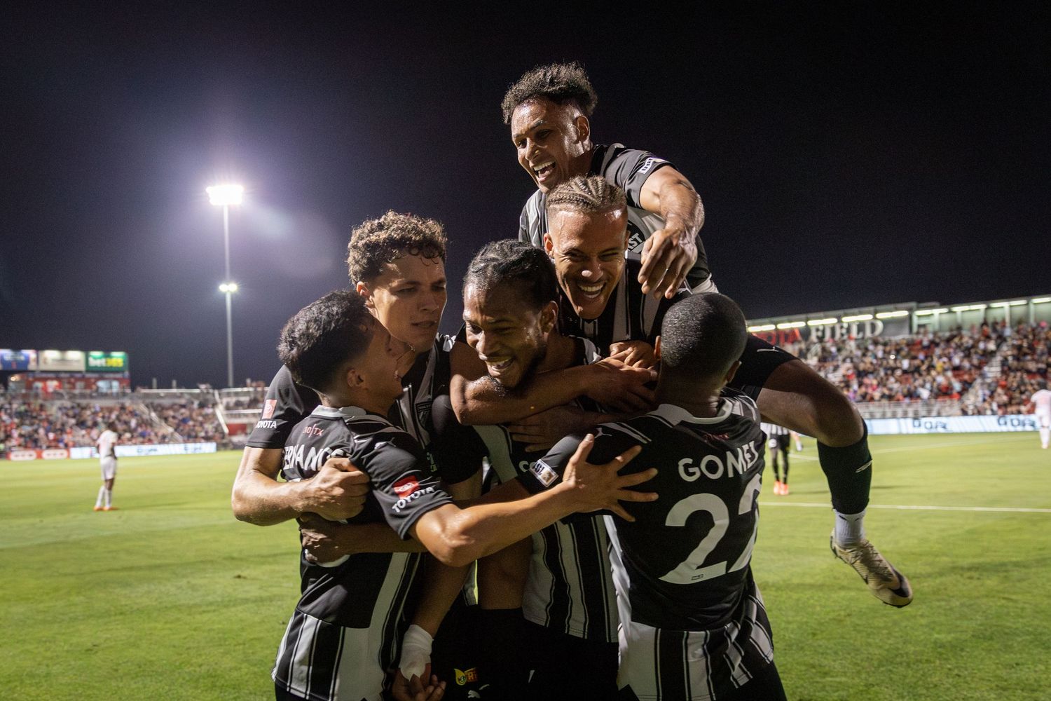 Soccer team in striped uniforms celebrates goal with group hug under stadium lights.