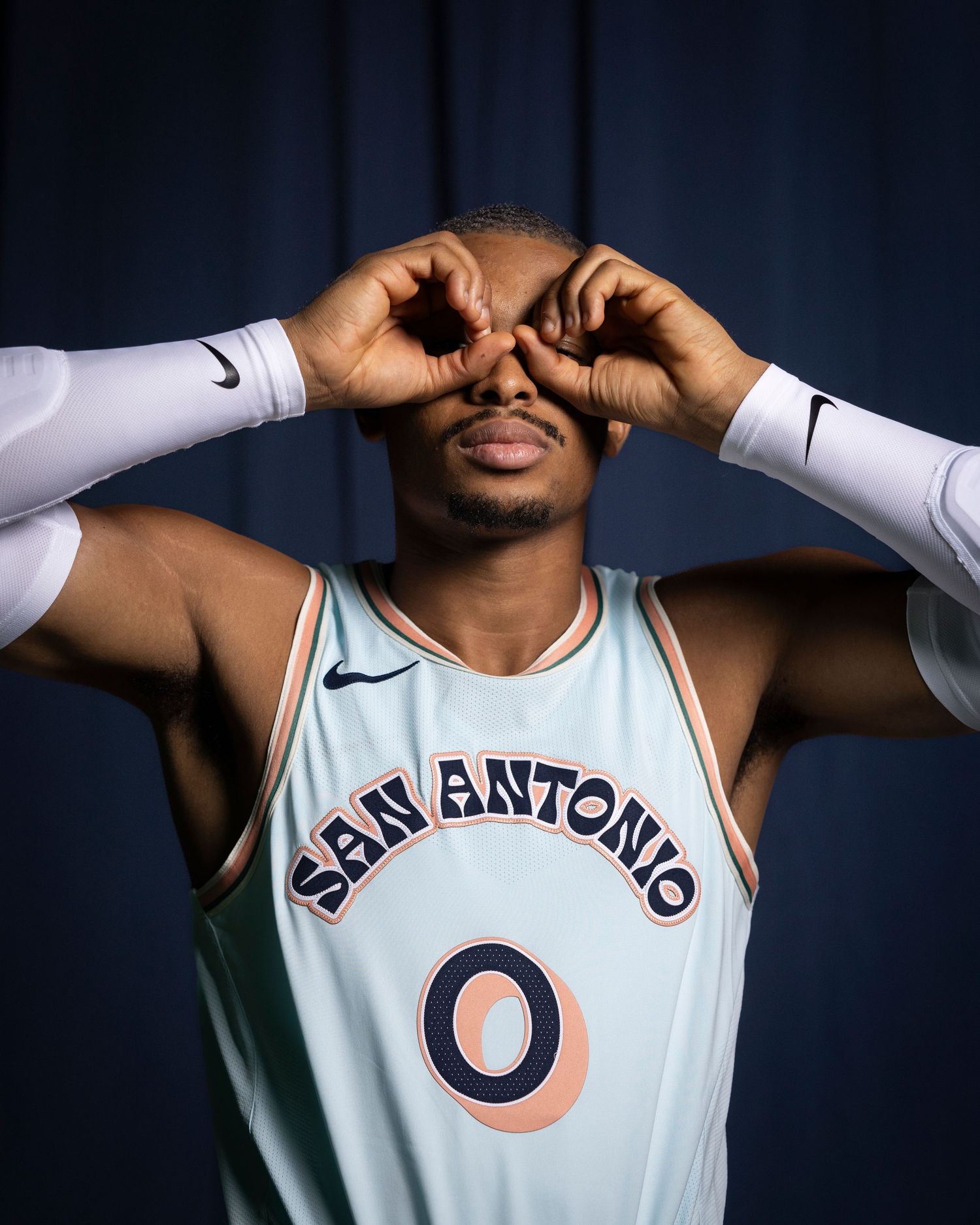 Basketball player in San Antonio jersey makes playful binoculars gesture with hands.