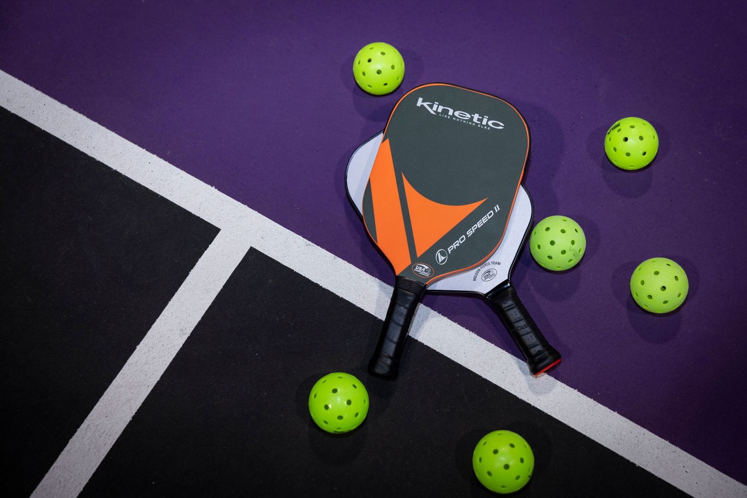 Black and orange pickleball paddle with neon yellow balls on purple court surface.