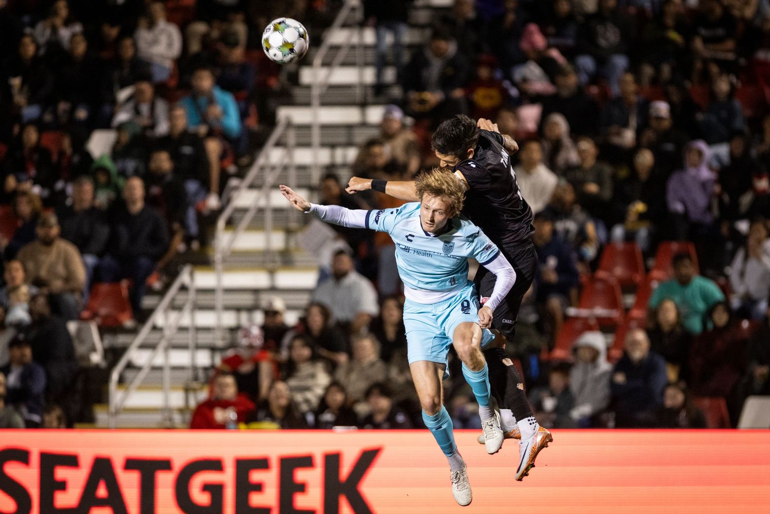 Soccer players compete for aerial ball during night match with stadium lighting.