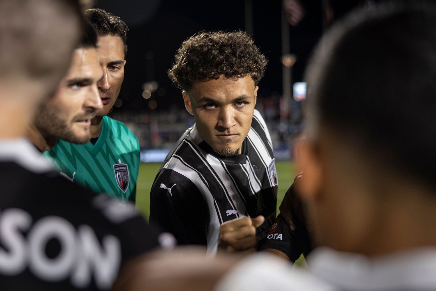 Team members gather for intense sideline discussion during nighttime soccer match.