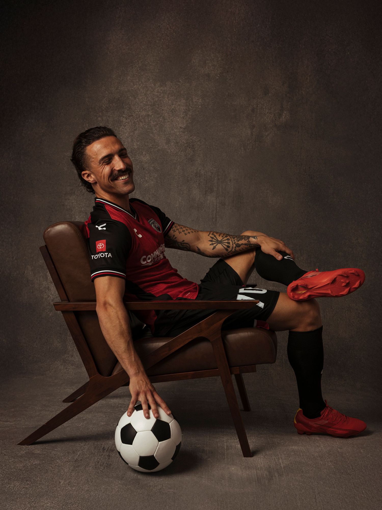 Soccer player in red and black kit sits relaxed in chair with ball, smiling, showing tattoos and red cleats.