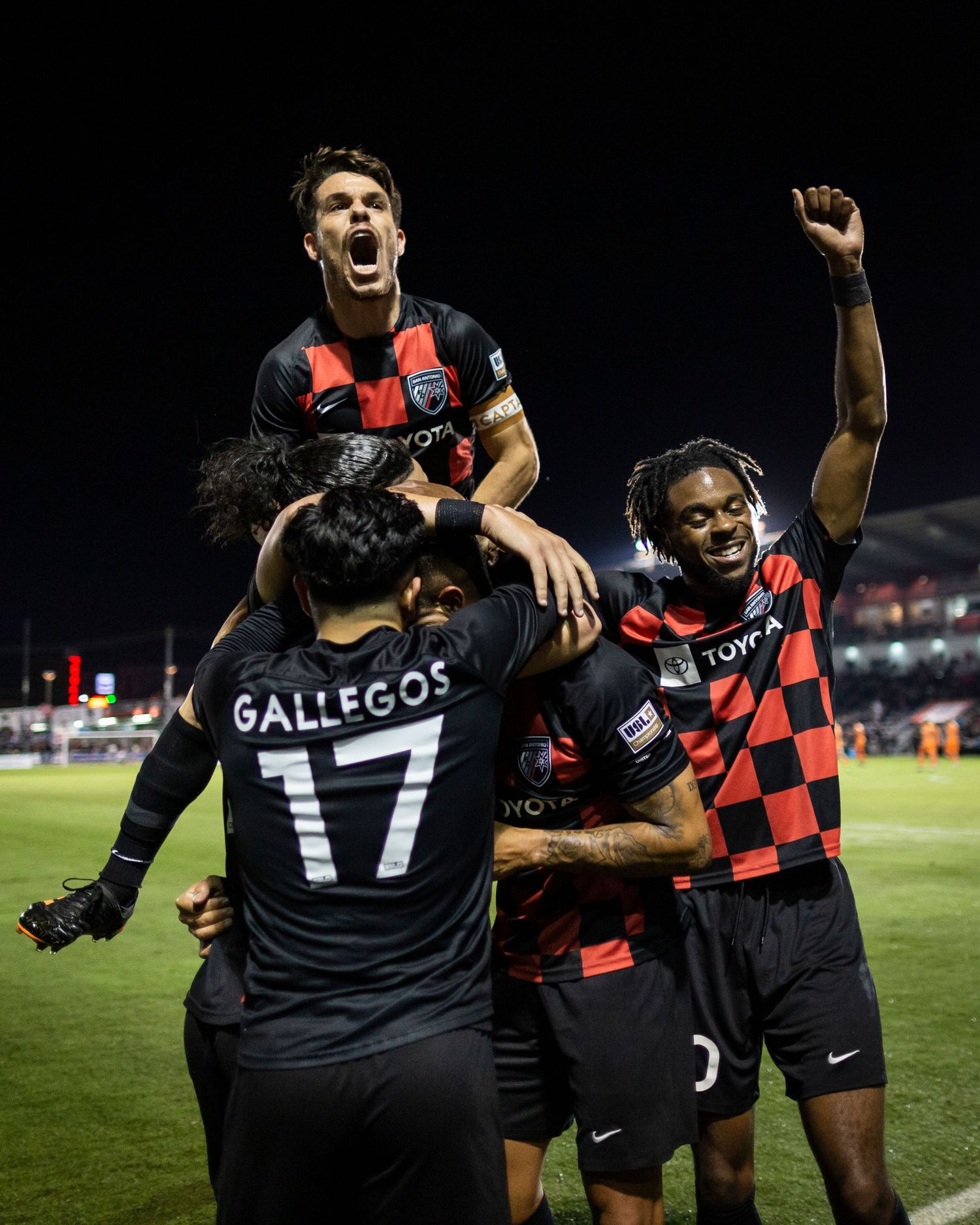 Rugby players celebrate together on the field during a nighttime match wearing red and black checkered uniforms.