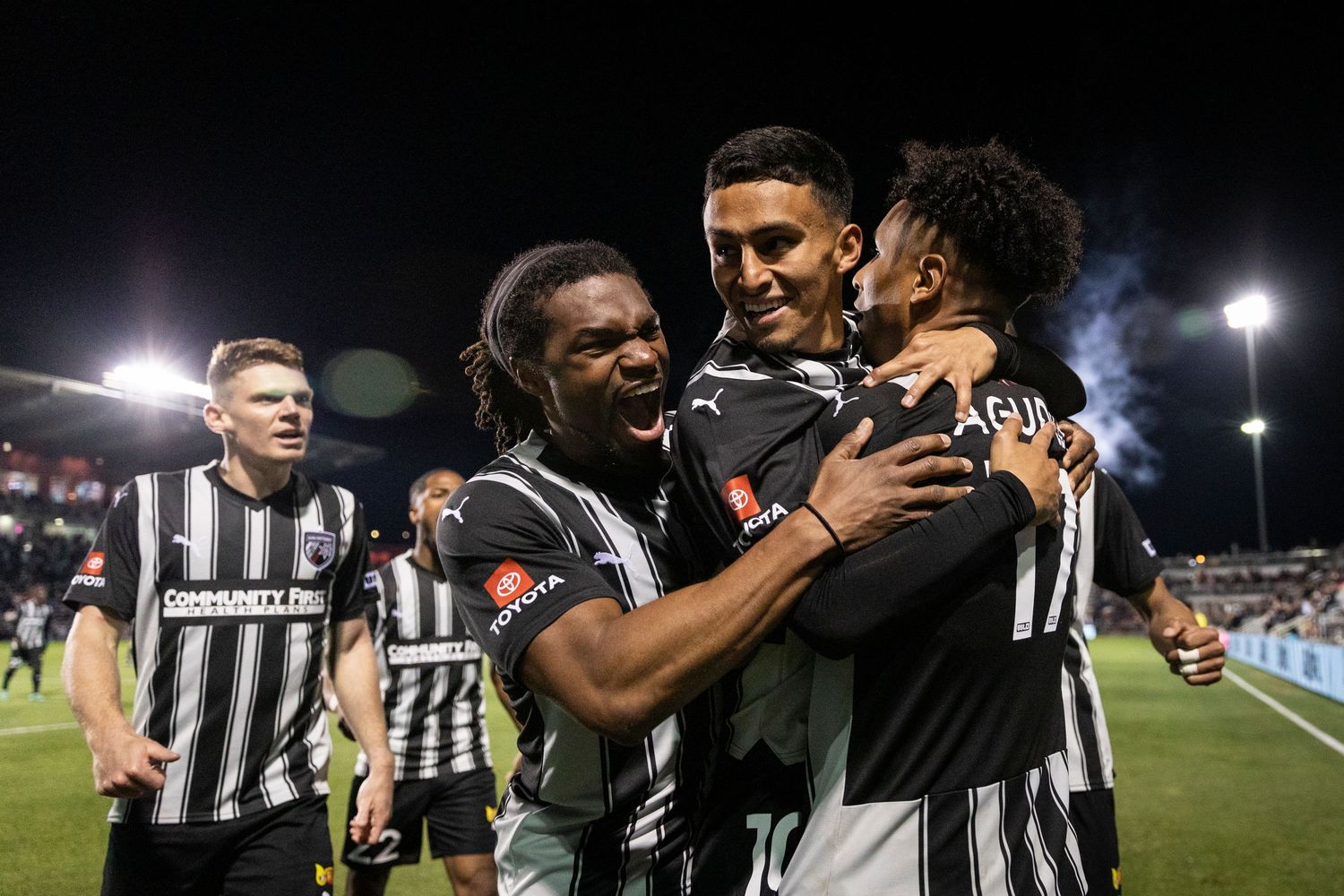 Soccer teammates celebrate together in black and white striped jerseys during nighttime match.